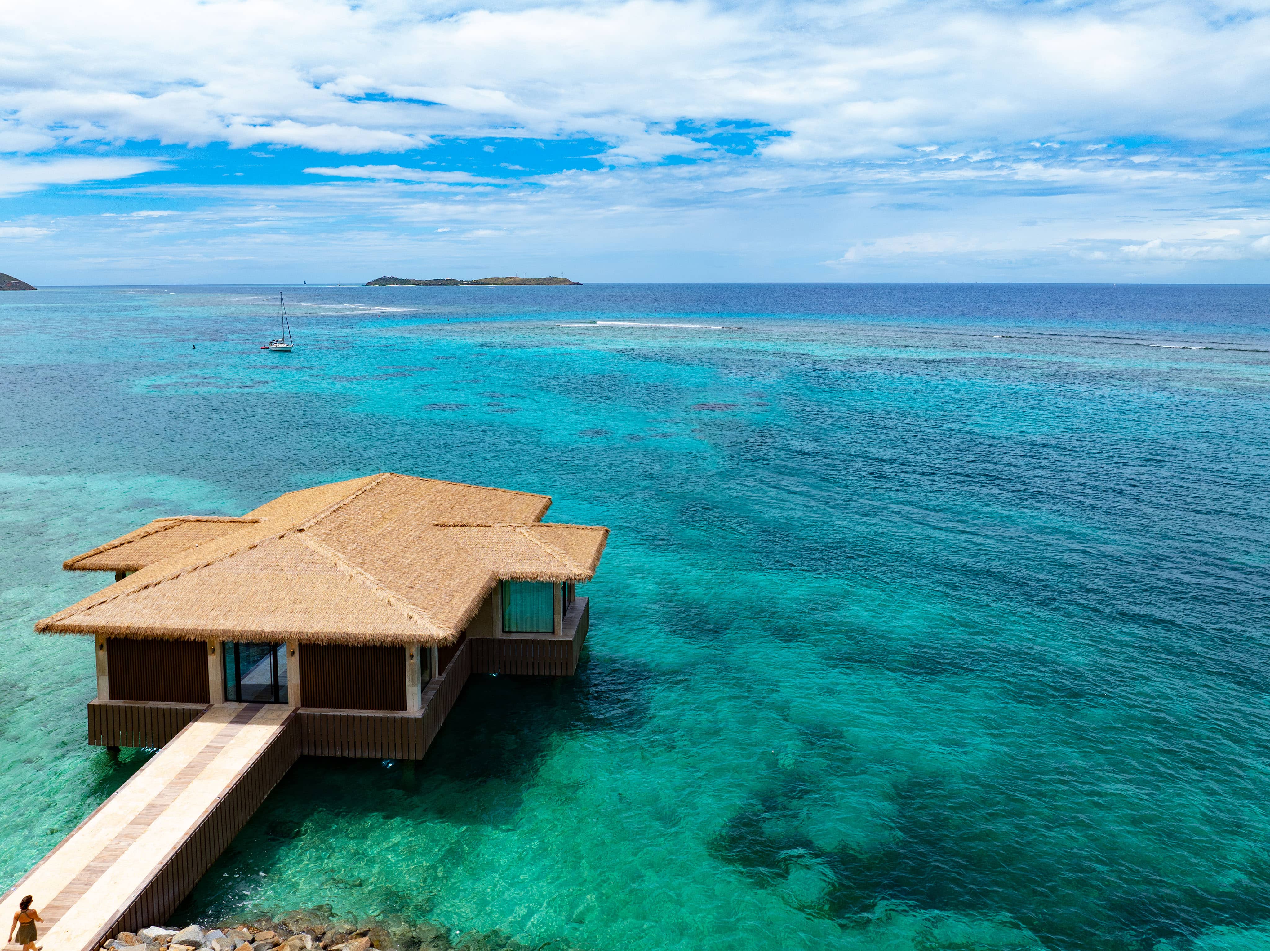 A thatched-roof overwater bungalow sits on turquoise, clear ocean water at Oil Nut Bay in the British Virgin Islands, with a wooden walkway leading to it, surrounded by coral reefs and distant islands under a partly cloudy sky.