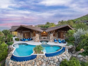 A luxurious villa with a unique, elevated design featuring stone and wood accents awaits at Oil Nut Bay. It overlooks a pool with curved edges surrounded by lush greenery. The sky is a blend of pink and purple hues, enhancing the scenic atmosphere of this British Virgin Islands luxury escape.
