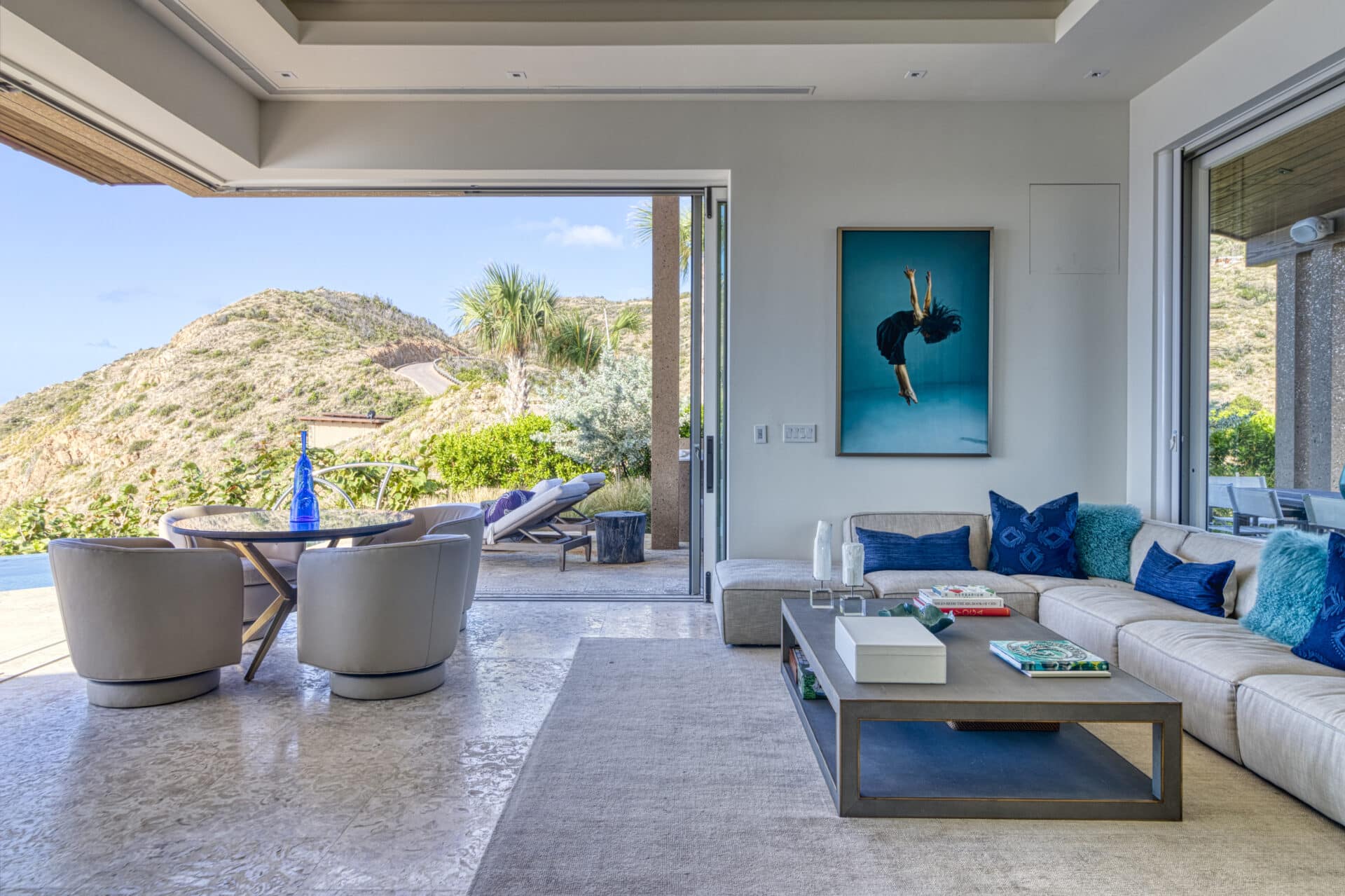 A spacious living room at Wild Side Villa boasts large glass doors opening to a scenic mountain view. The room features a beige sectional sofa with blue cushions, a modern rectangular coffee table, a round dining table with chairs, and a wall art piece depicting an upside-down diver.