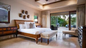 A spacious and luxurious bedroom in this $15.9M British Virgin Islands home features a wooden bed with white linens, an upholstered bench at the foot, and a lounge chair. Wooden furniture accents the room, while large sliding glass doors open to a lush garden with palm trees and outdoor seating.