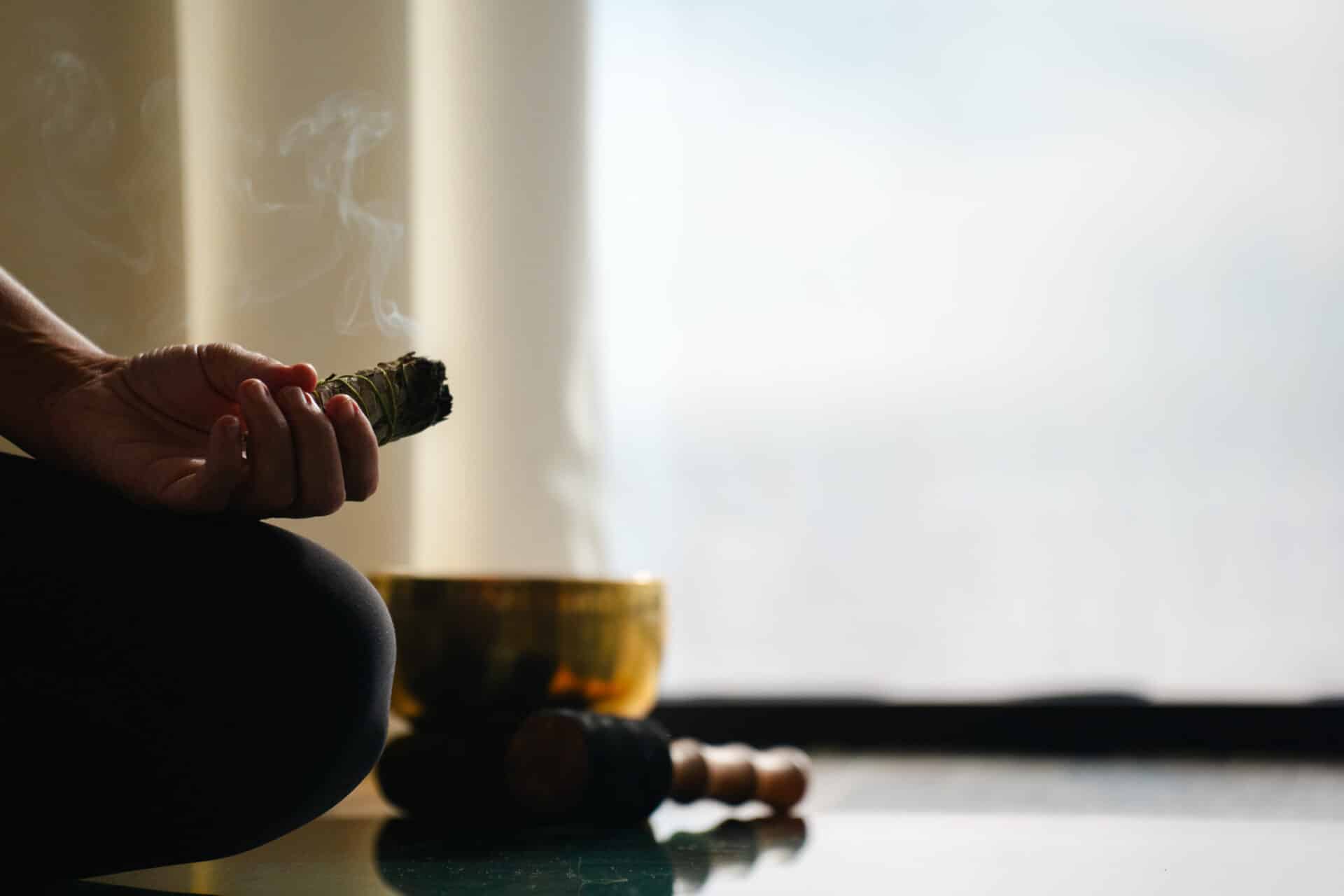 A person sits cross-legged indoors at Oil Nut Bay spa, holding a smoking bundle of sage. A singing bowl and mallet rest nearby, suggesting a tranquil scene of meditation or spiritual practice in soft, natural light.