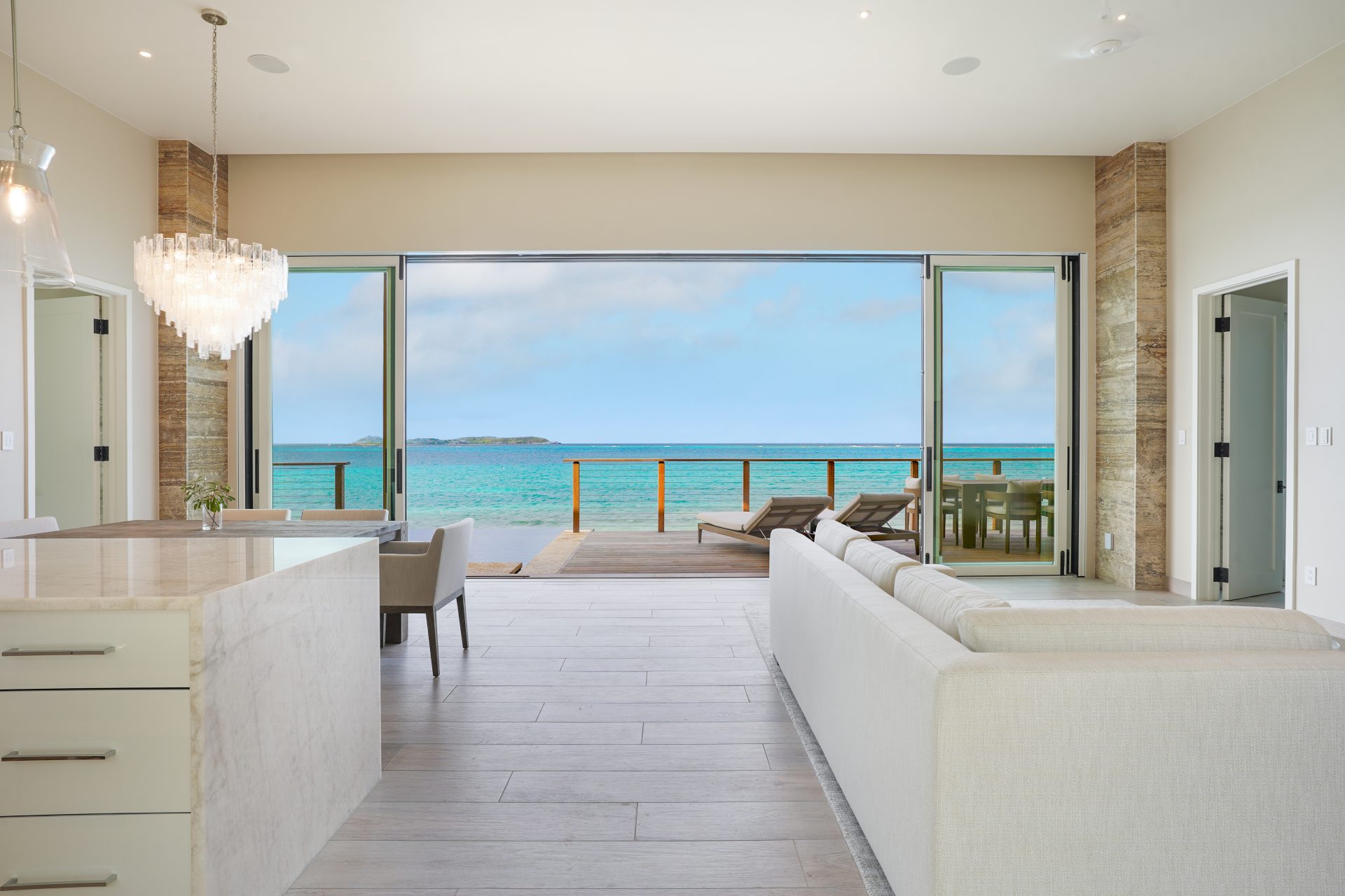 A modern, open-concept living space in the Ocean Villa features a light-colored sofa and a large kitchen island with quartz countertops. Floor-to-ceiling glass doors open to a patio with lounge chairs, offering a stunning ocean view and seamless transition between indoor and outdoor living.