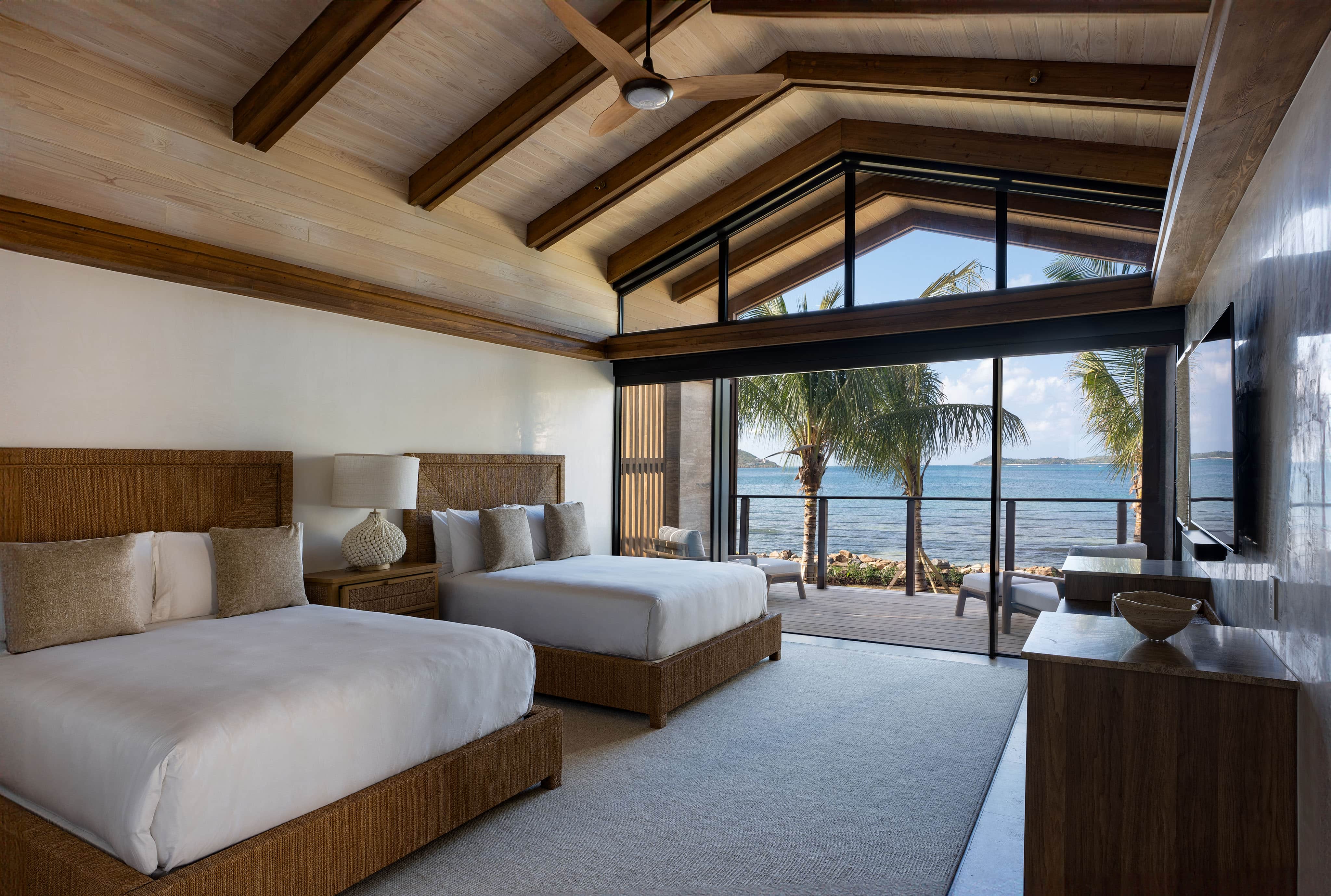 This Ocean Villa boasts a luxurious bedroom with two double beds, wooden ceiling beams, and large windows. Step onto the balcony to enjoy views of palm trees and the ocean. Neutral tones and warm lighting in this OV1 create a cozy atmosphere.