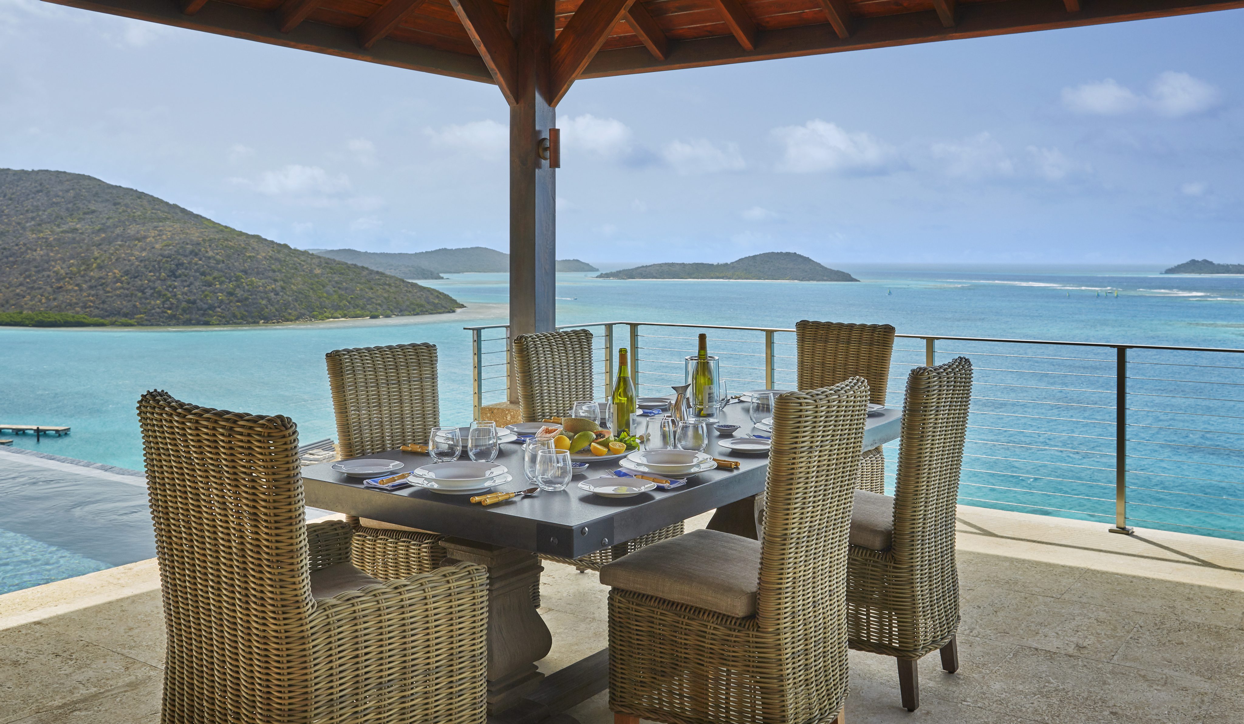 A dining table set with plates, glasses, and wine bottles is positioned under a shaded pergola on a patio at the Reef House, overlooking a serene turquoise ocean and lush green islands. The scene exudes a tranquil, luxurious outdoor dining experience with stunning coastal views.