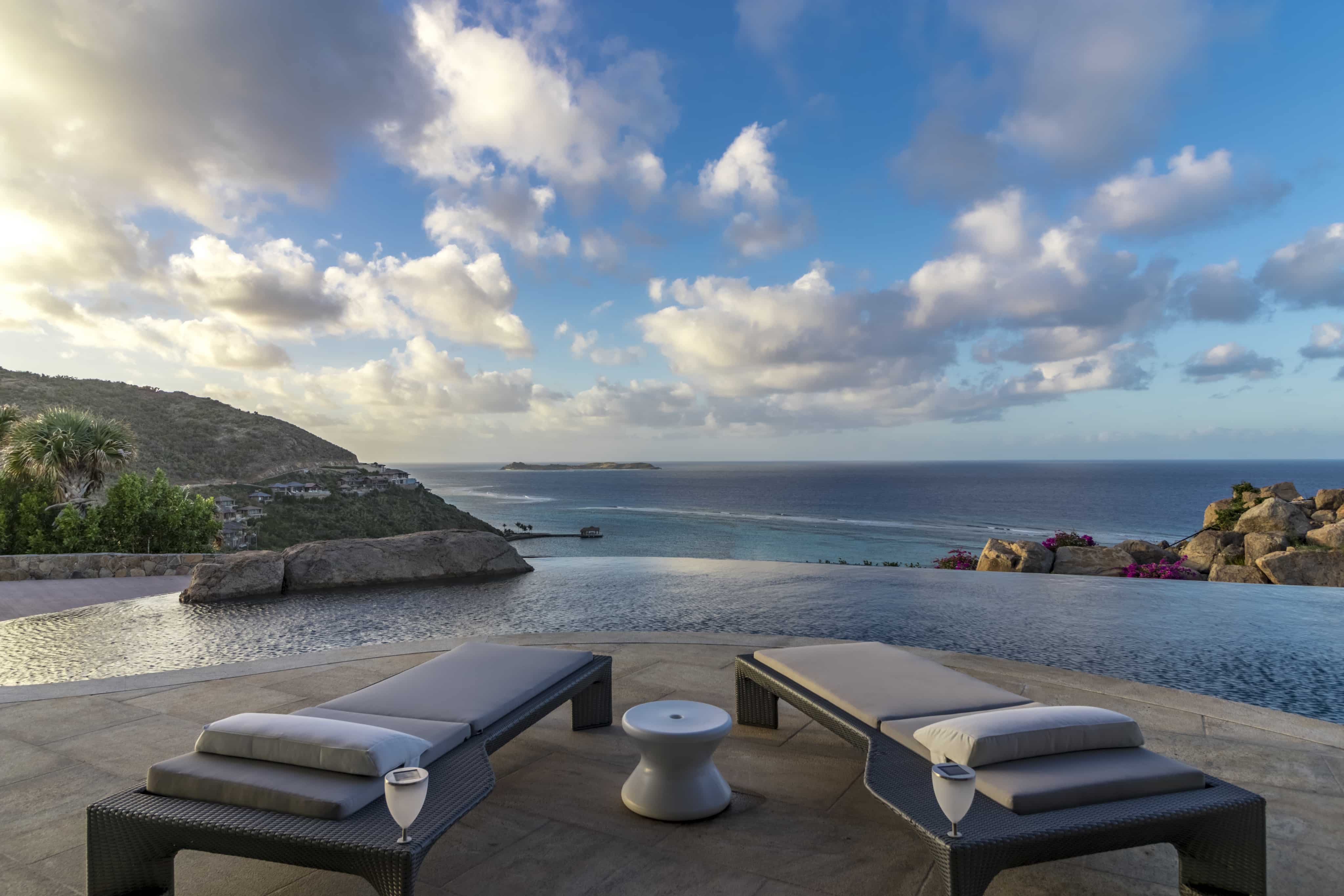 An infinity pool at the villa overlooks a scenic view of the ocean and distant islands under a partly cloudy sky. Two lounge chairs and a small table rest on the pool deck, with glasses placed on the table. Rocky hillsides and foliage frame the tranquil scene, creating an idyllic halo of serenity.