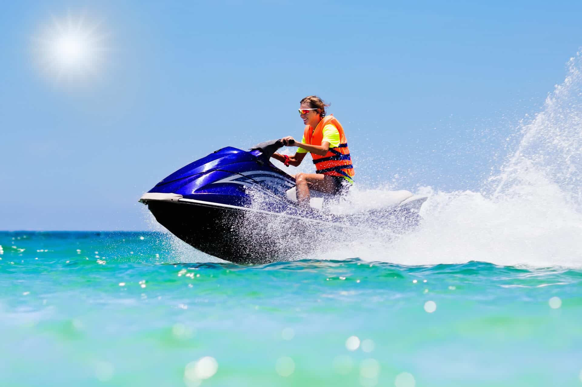 A person wearing an orange life jacket and sunglasses enjoys jetskiing on a blue jet ski across clear, turquoise waters under a sunny blue sky. The jet ski creates a splash of water as it moves, and the scene is bright and energetic.