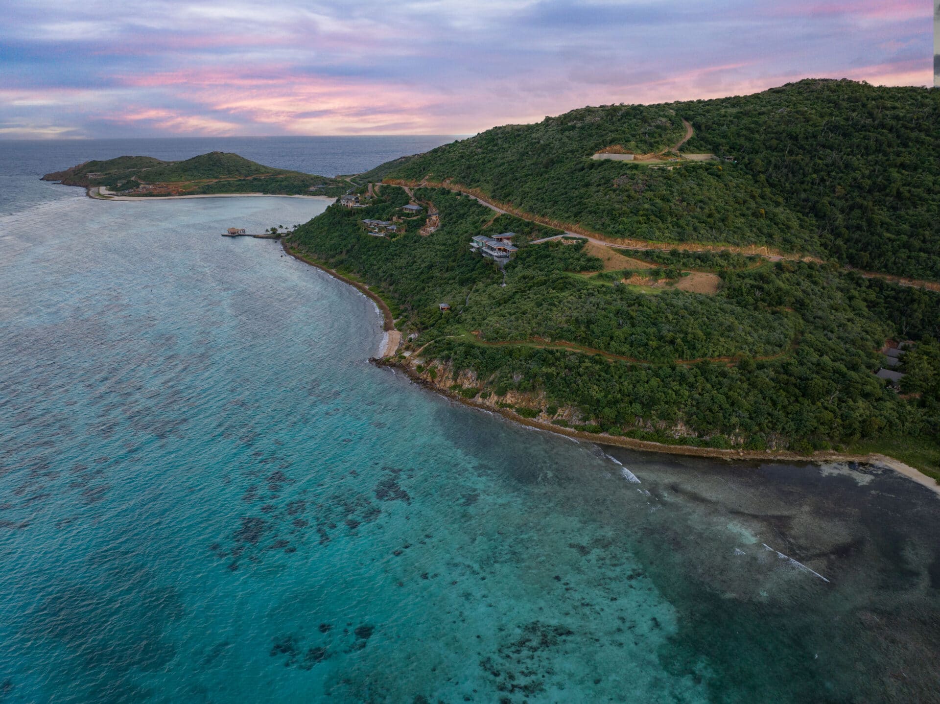 Aerial view of a coastal landscape with lush green hills and clear turquoise waters. The E15 road elegantly winds along the coastline. The sky, a mix of pink and blue hues, suggests either sunrise or sunset. Estate homesites dot the area, and small islands are visible in the distance.