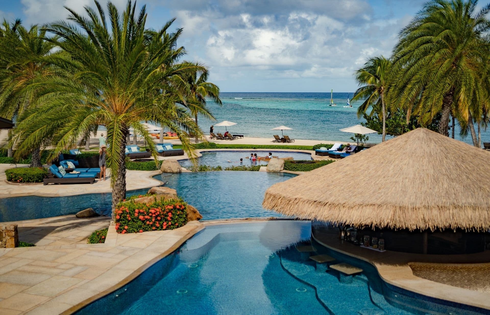 Tropical resort in Oil Nut Bay, British Virgin Islands, featuring a luxurious pool area with sun loungers, a thatched-roof bar, and palm trees. Beyond the pool, guests relax on a sandy beach while enjoying the clear blue ocean. A few small boats and lush landscaping complete this luxury sanctuary.