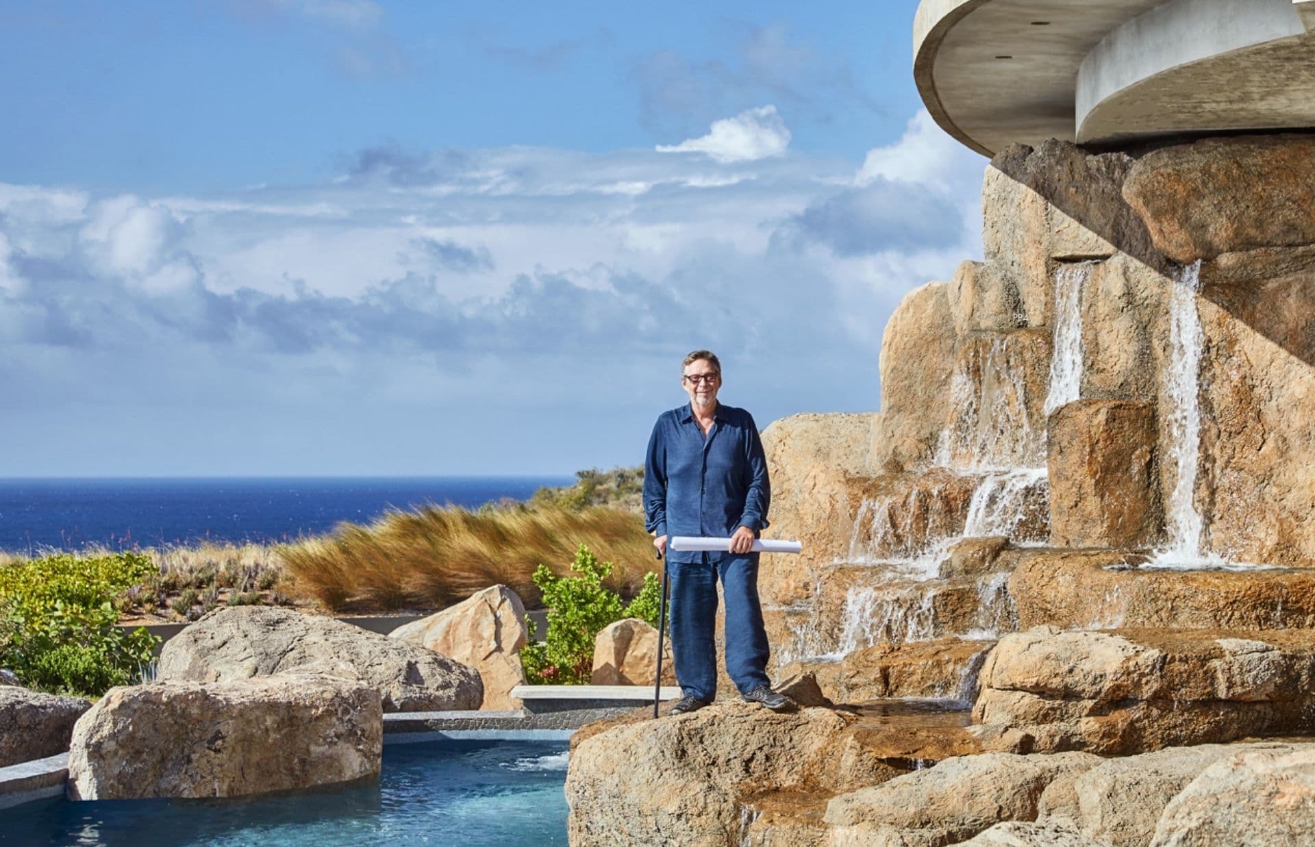 David V. Johnson stands on a rocky ledge overlooking a pool with cascading water. Dressed in a blue shirt and jeans, he holds a rolled paper, gazing at the beautiful ocean view and partially cloudy sky in BVI's serene Oil Nut Bay. The setting is serene and picturesque.