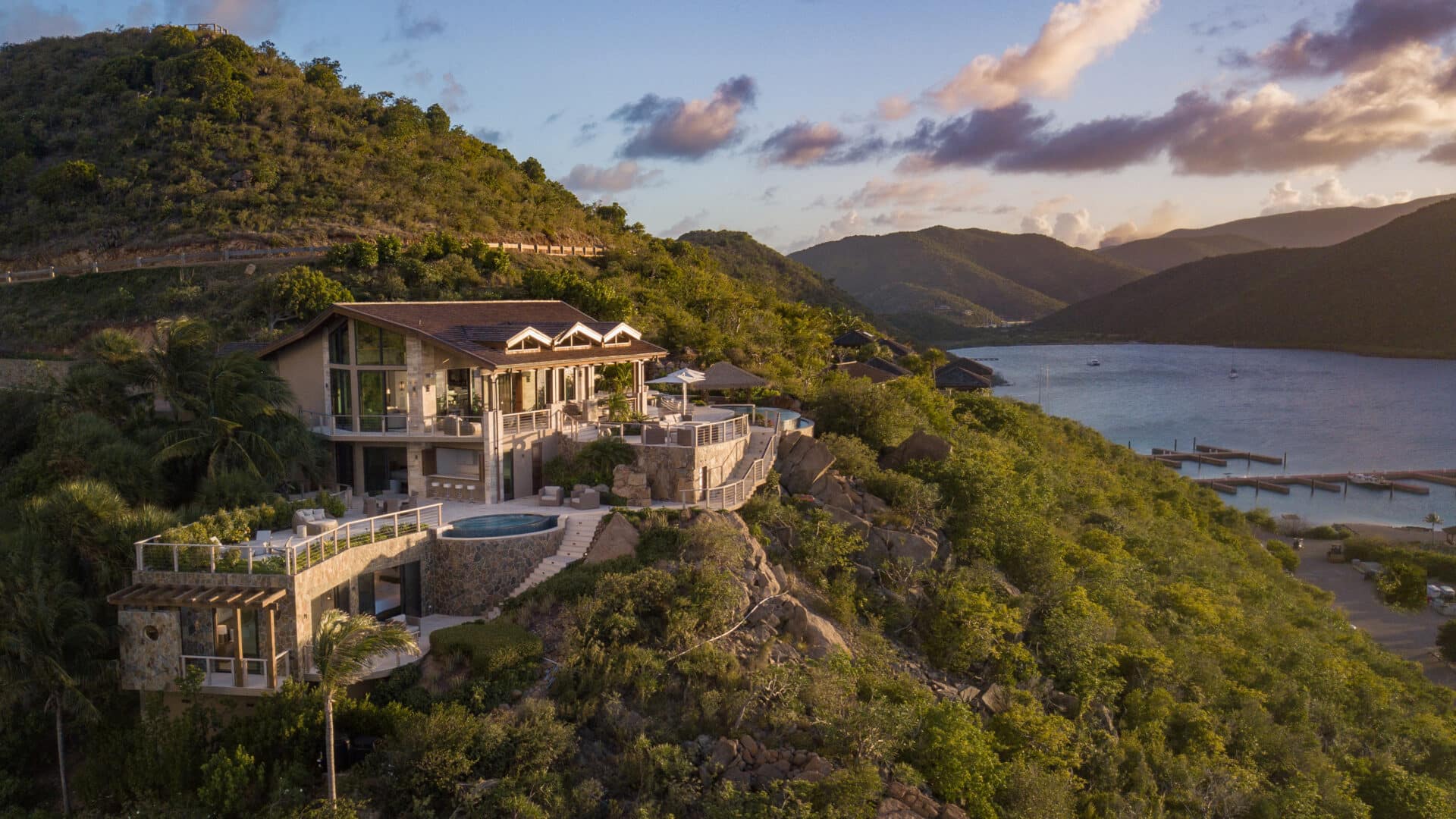 A luxurious cliffside villa with a pool, surrounded by lush greenery, overlooks a serene bay in the British Virgin Islands. The sky is painted with soft clouds during a warm sunset, and boats are visible in the water below. Rolling hills frame this picturesque scene.