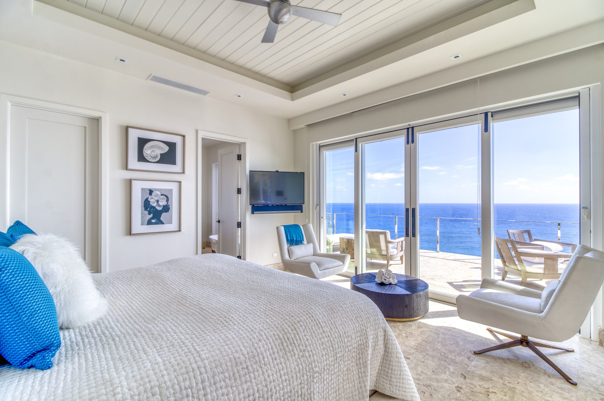 A bright and modern bedroom at Wild Side Villa featuring a large bed with white and blue bedding, a wall-mounted TV, and framed art. Floor-to-ceiling windows and sliding glass doors lead to a balcony with a seating area, offering a stunning view of the ocean.