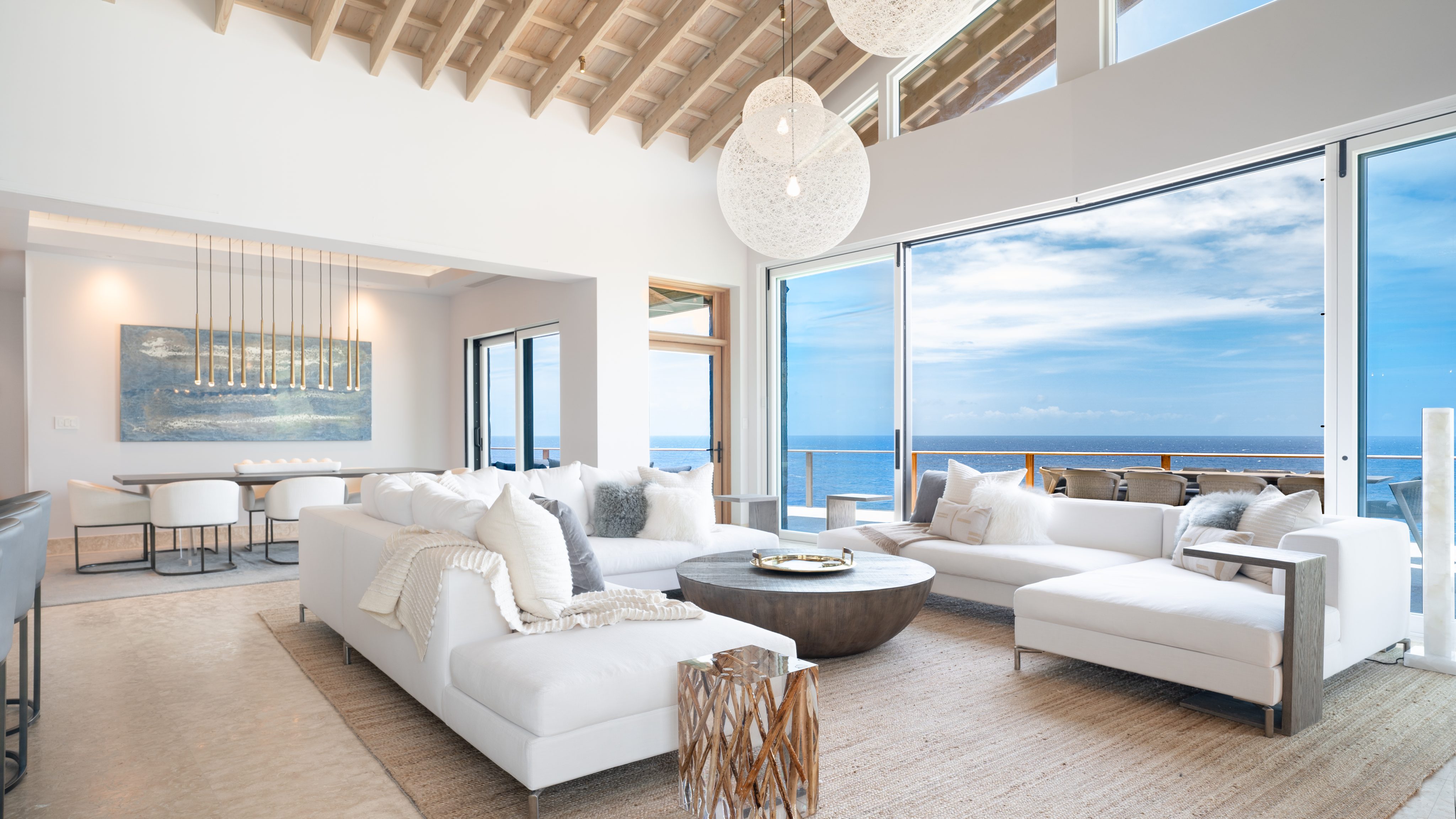 A spacious, modern living room in the villa with large windows showcasing an ocean view. The room features white sofas, a round coffee table, and contemporary hanging light fixtures. A dining area with a long table and chairs is visible in the background. Natural light fills the space, perfect for spotting an occasional hummingbird.