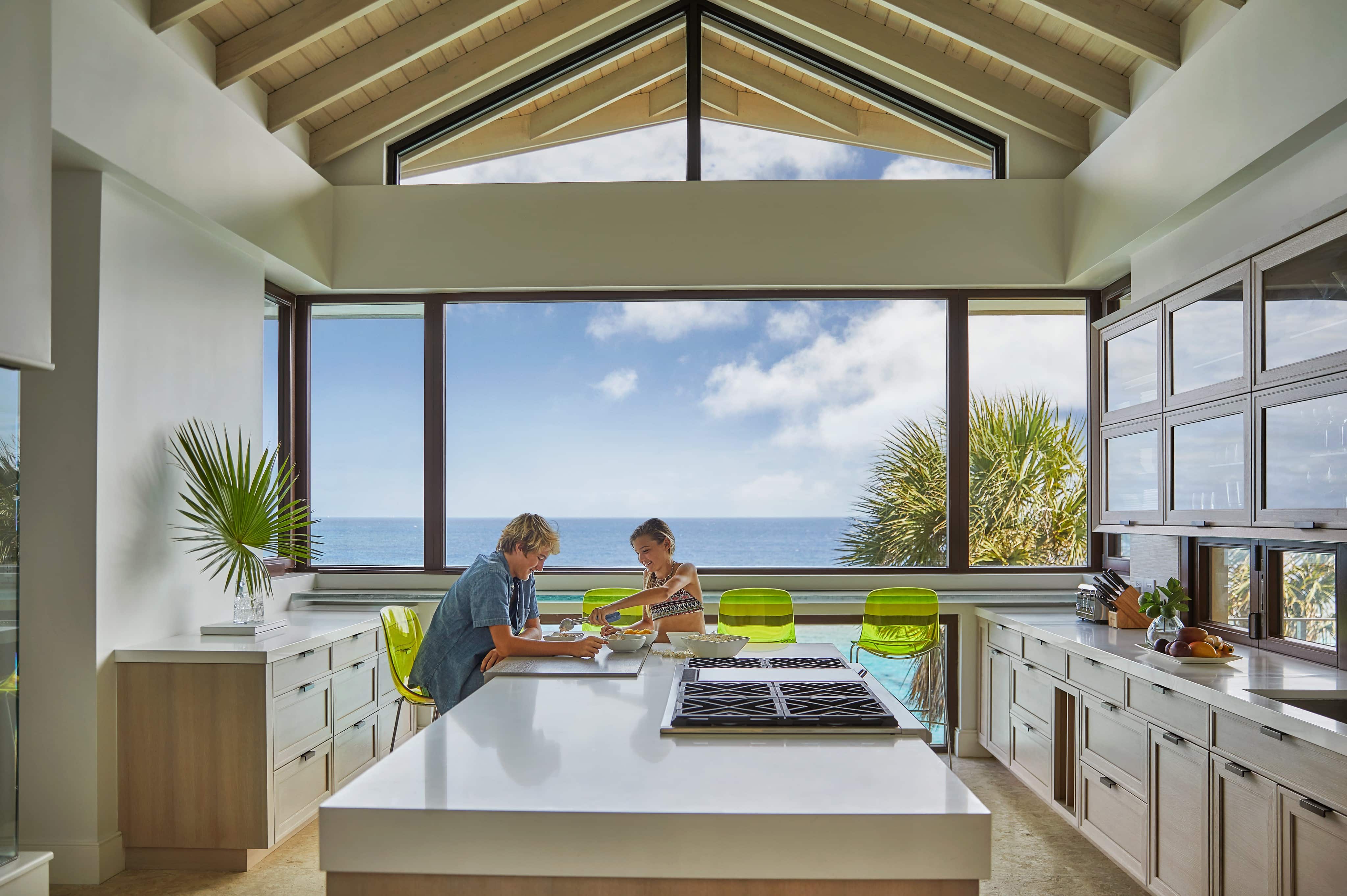 A spacious, modern kitchen with large windows overlooking the ocean. Two people sit at a white island counter; one is writing, and the other is observing. Bright green chairs add a pop of color, and a palm frond decorates the counter. About them, the sky is clear and sunny outside.