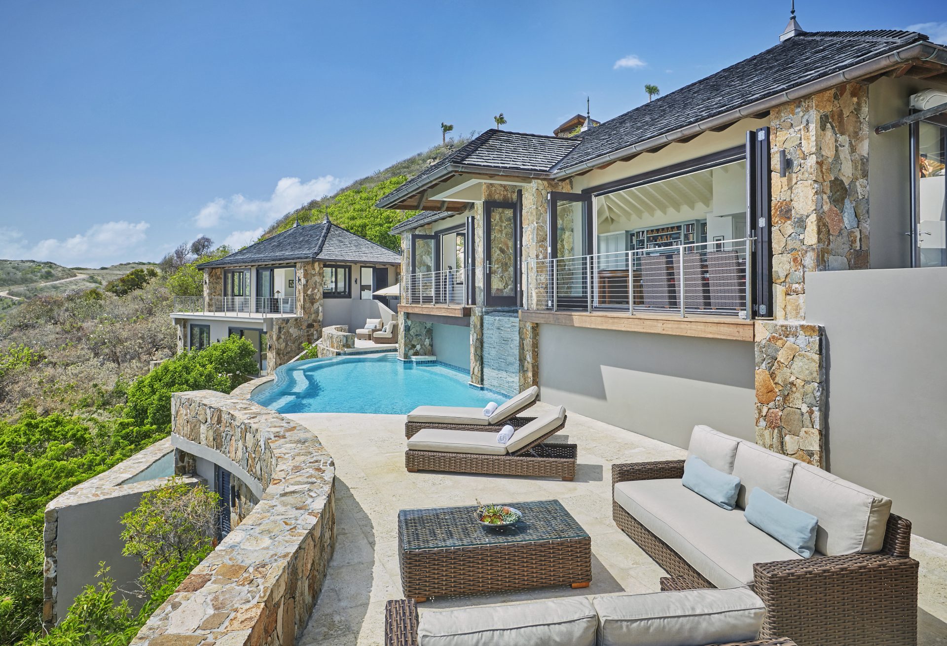 Cooke Lookout Villa is a luxurious hillside retreat with stone walls and an infinity pool that overlooks a lush green valley. The outdoor area includes sun loungers, a wicker coffee table, and a cushioned patio set under a clear blue sky. Adjoining rooms have large windows and sliding doors for breathtaking views.