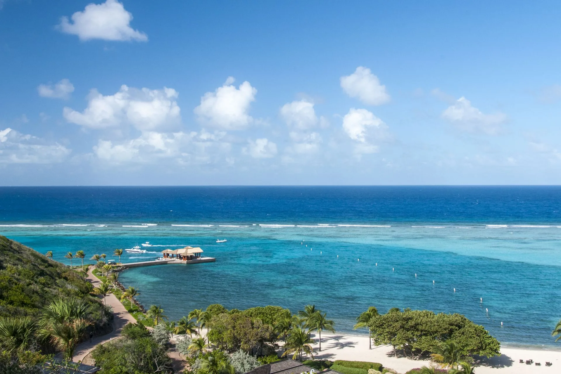 Experience a scenic view of a tropical coastline at one of our Luxurious Caribbean Resorts, featuring clear blue waters, a small dock extending into the sea, lush green palm trees, and a sandy beach. Reopening this fall, with the calm ocean meeting a sky dotted with white clouds.