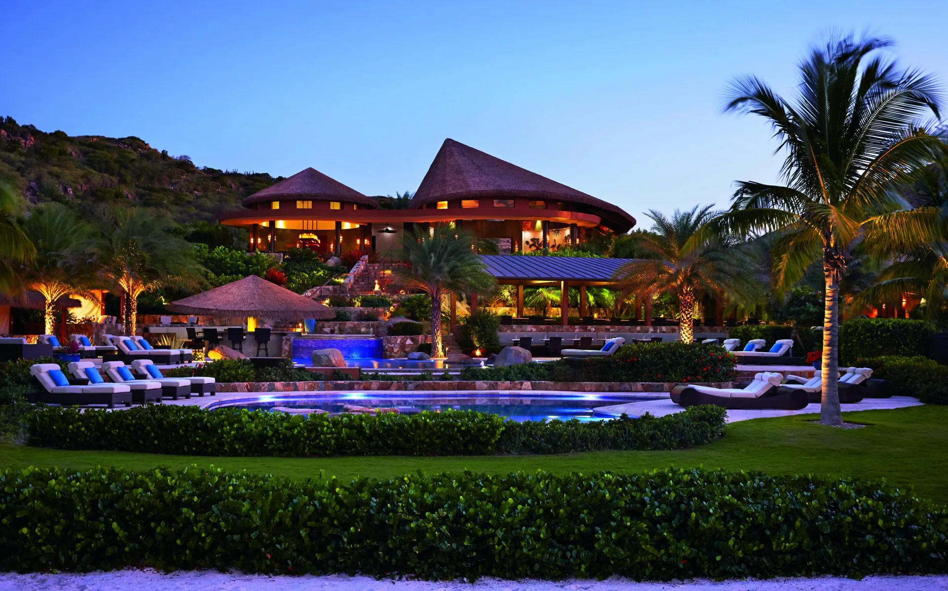 An upscale resort at twilight featuring a thatch-roofed building with warm lighting, a multi-tiered swimming pool, sun loungers, and lush greenery. This one-of-a-kind oasis is adorned with palm trees under a clear evening sky.