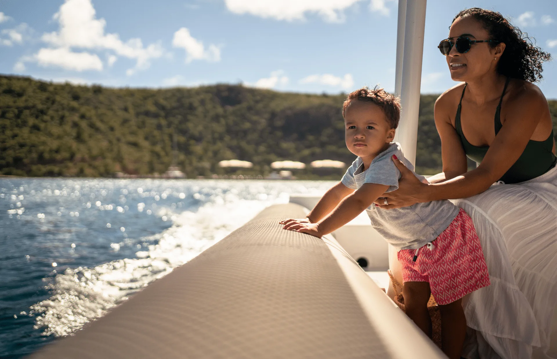 A woman and a young child are on a boat, leaning against the edge and looking out over the water. The woman wears sunglasses and holds the child securely. The background features a scenic view of the best Caribbean waters and green hills under a sunny sky.