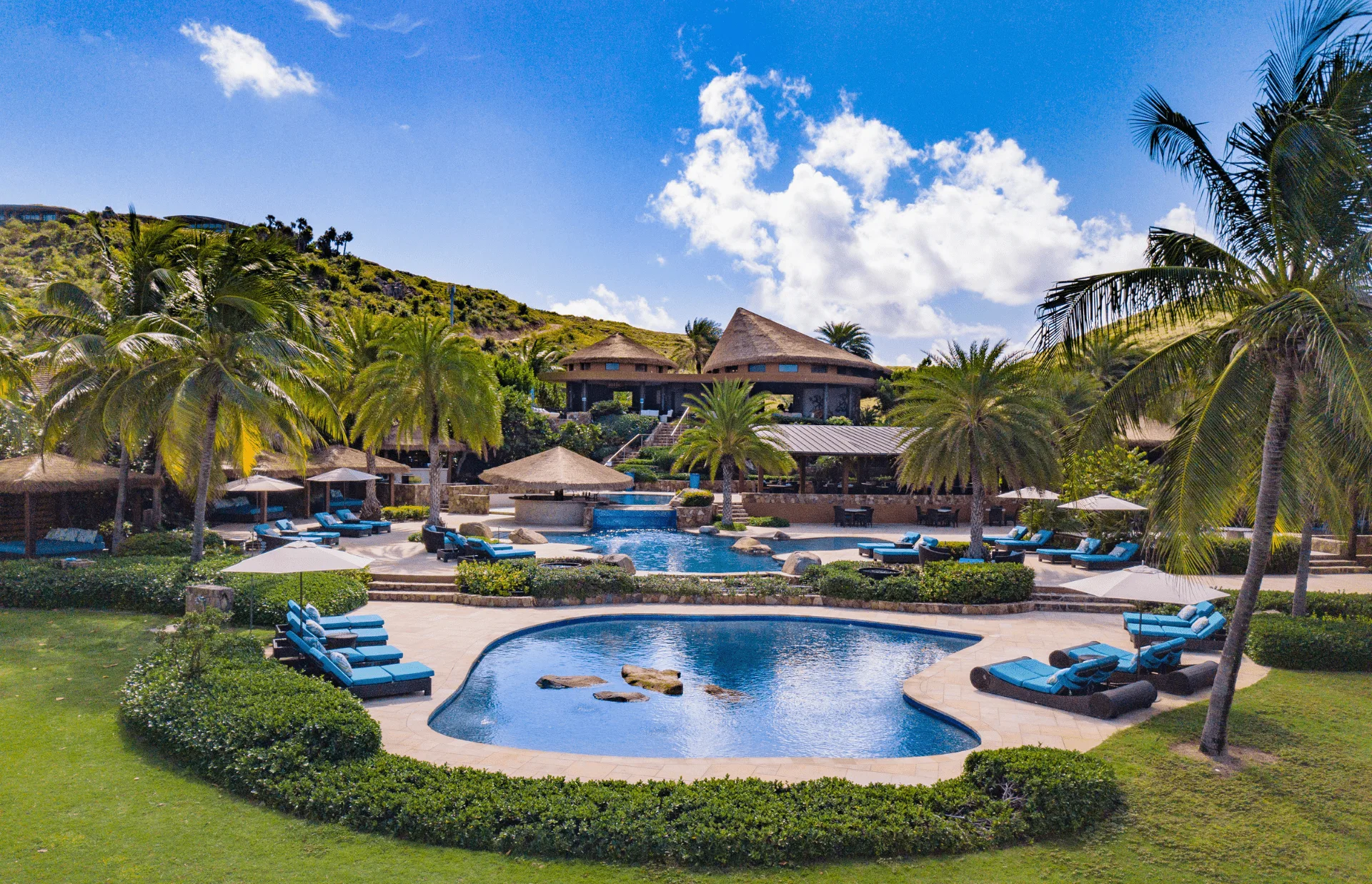 A luxurious resort perfect for Valentine's Day getaways features a sprawling pool area with multiple connected pools, surrounded by lush greenery and tall palm trees. Lounge chairs with blue cushions line the poolside, while thatched-roof huts and a large villa make for the ultimate romantic escape under a bright blue sky.