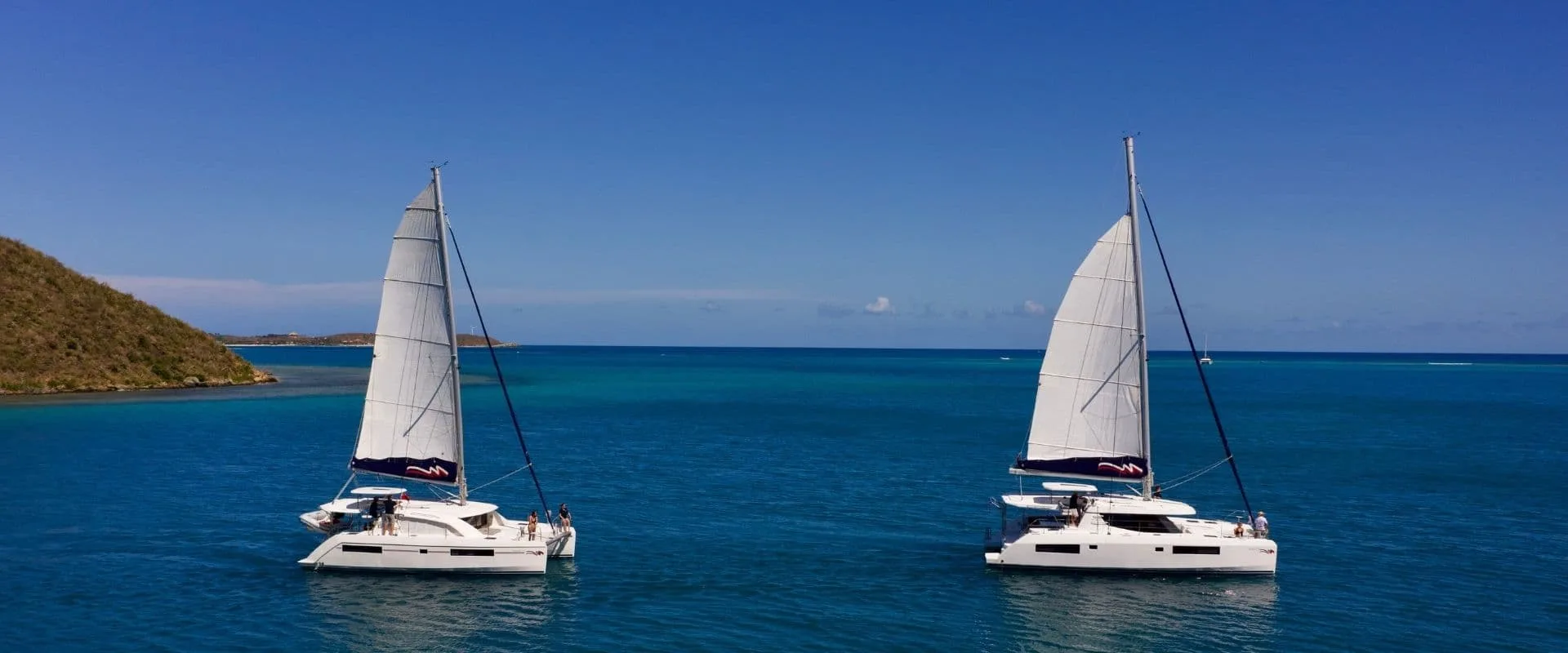 Two white catamarans with raised sails float on a calm, deep blue sea under a clear sky. They are sailing side by side near a coastline featuring a slope covered in green vegetation on the left. The horizon separates the sea from the sky in the background, making it an ideal scene for travelers to the BVI.