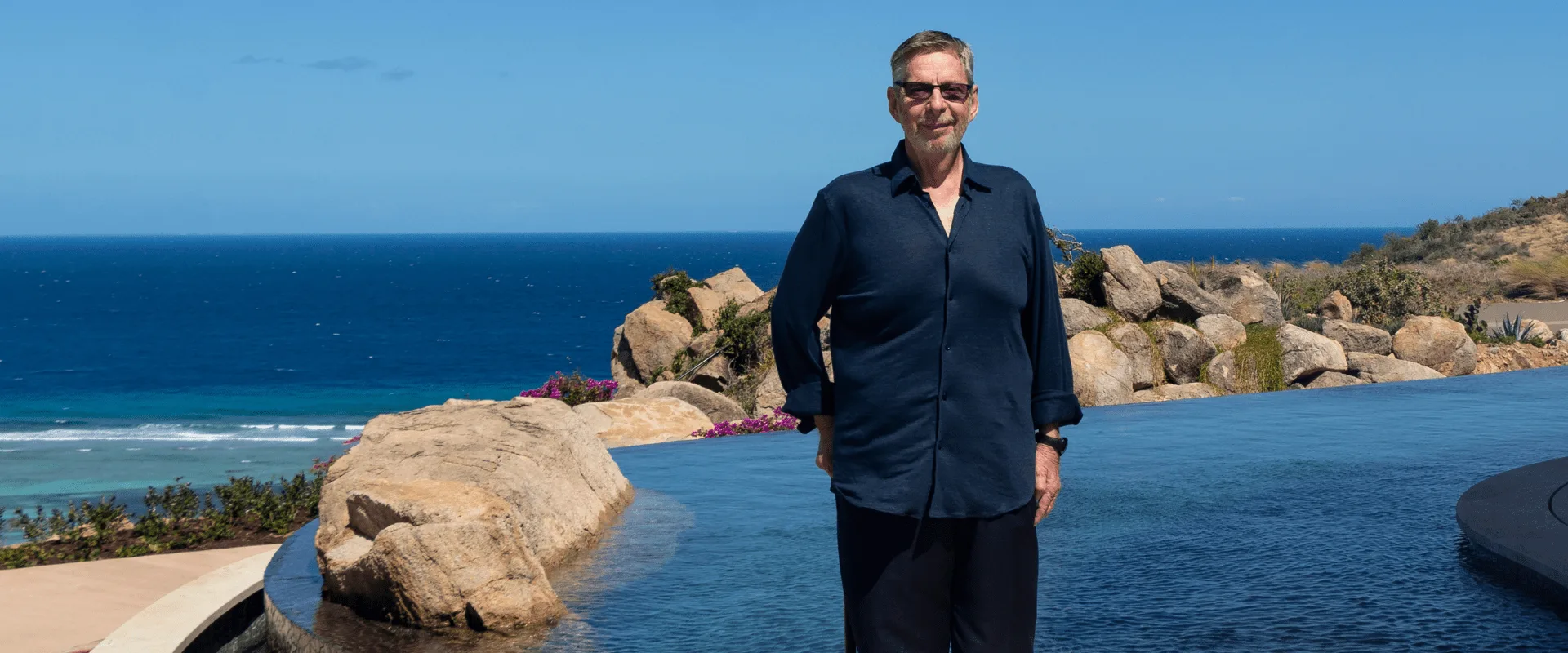 A man wearing sunglasses and a dark button-up shirt stands by an infinity pool, with the ocean and a rocky coastline in the background on a clear day, embodying the relaxed sophistication of David Johnson, a notable figure in the Caribbean yachting community.