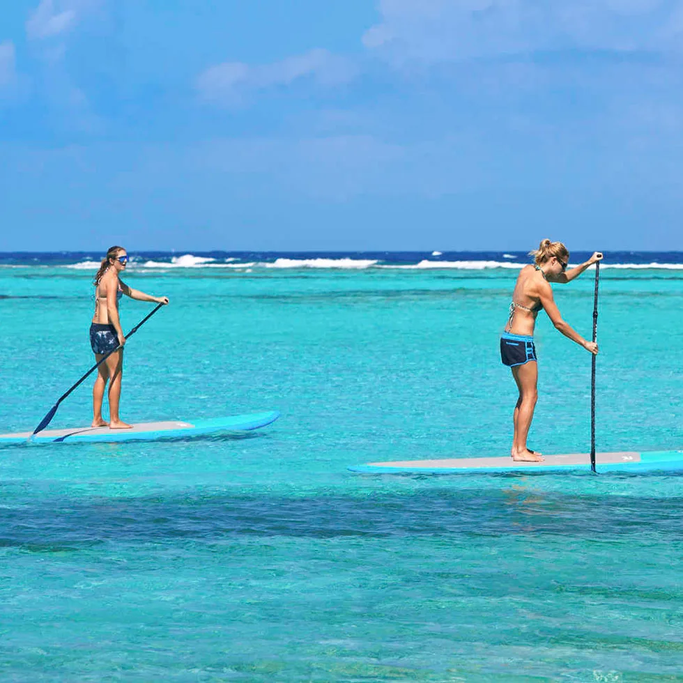 Two people paddleboarding on clear turquoise water at Oil Nut Bay. Both are standing on separate boards in swimwear, using long paddles to navigate. The horizon in the background shows a deep blue ocean and some light waves, with a bright blue sky above.