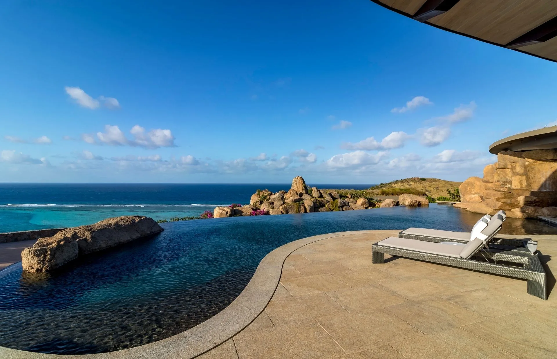 Infinity pool with a breathtaking view overlooking a rocky coastline and the ocean, under a clear blue sky. A white lounge chair is placed beside the pool on a tiled deck, creating a serene and luxurious atmosphere perfect for those seeking luxurious homes.