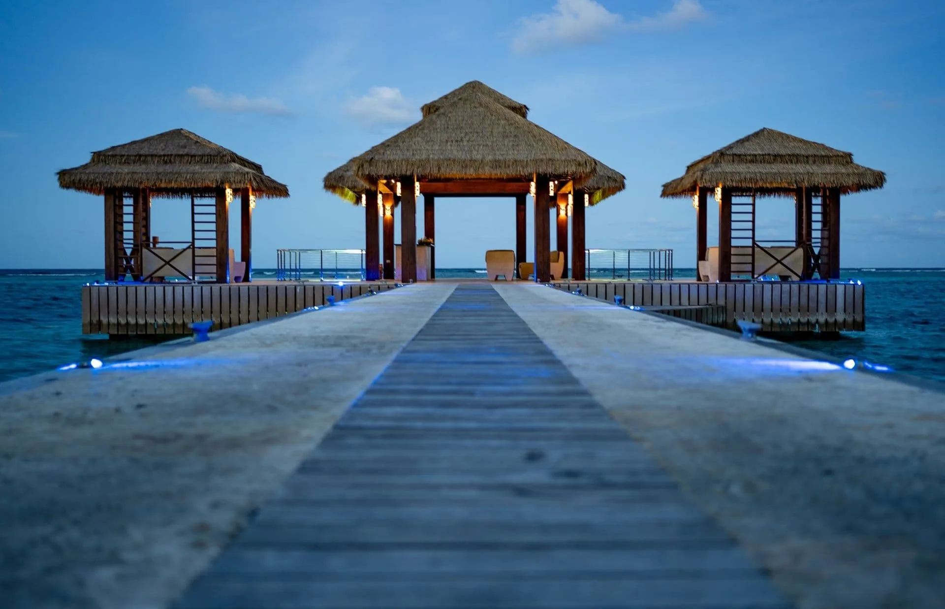 A serene view of a wooden walkway leading to three thatched-roof cabanas over the water at Oil Nut Bay, illuminated by soft blue lights at dusk. The peaceful ocean and a clear sky provide a calm backdrop, exemplifying true Location Luxe.