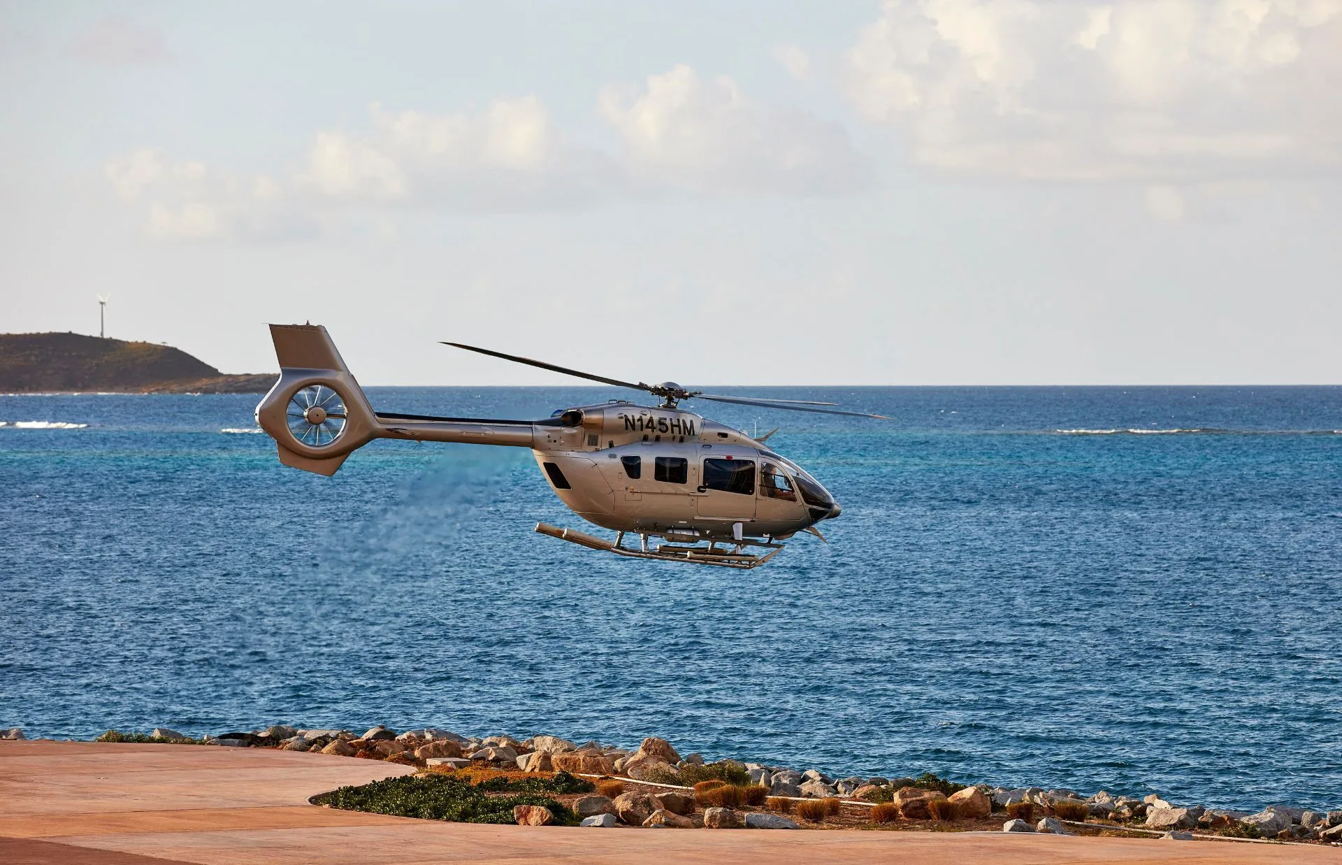 A gray helicopter hovers over a coastal area with a rocky shoreline. The background features a vast expanse of calm blue ocean and a partly cloudy sky. In the distance, a piece of land with a wind turbine is visible.