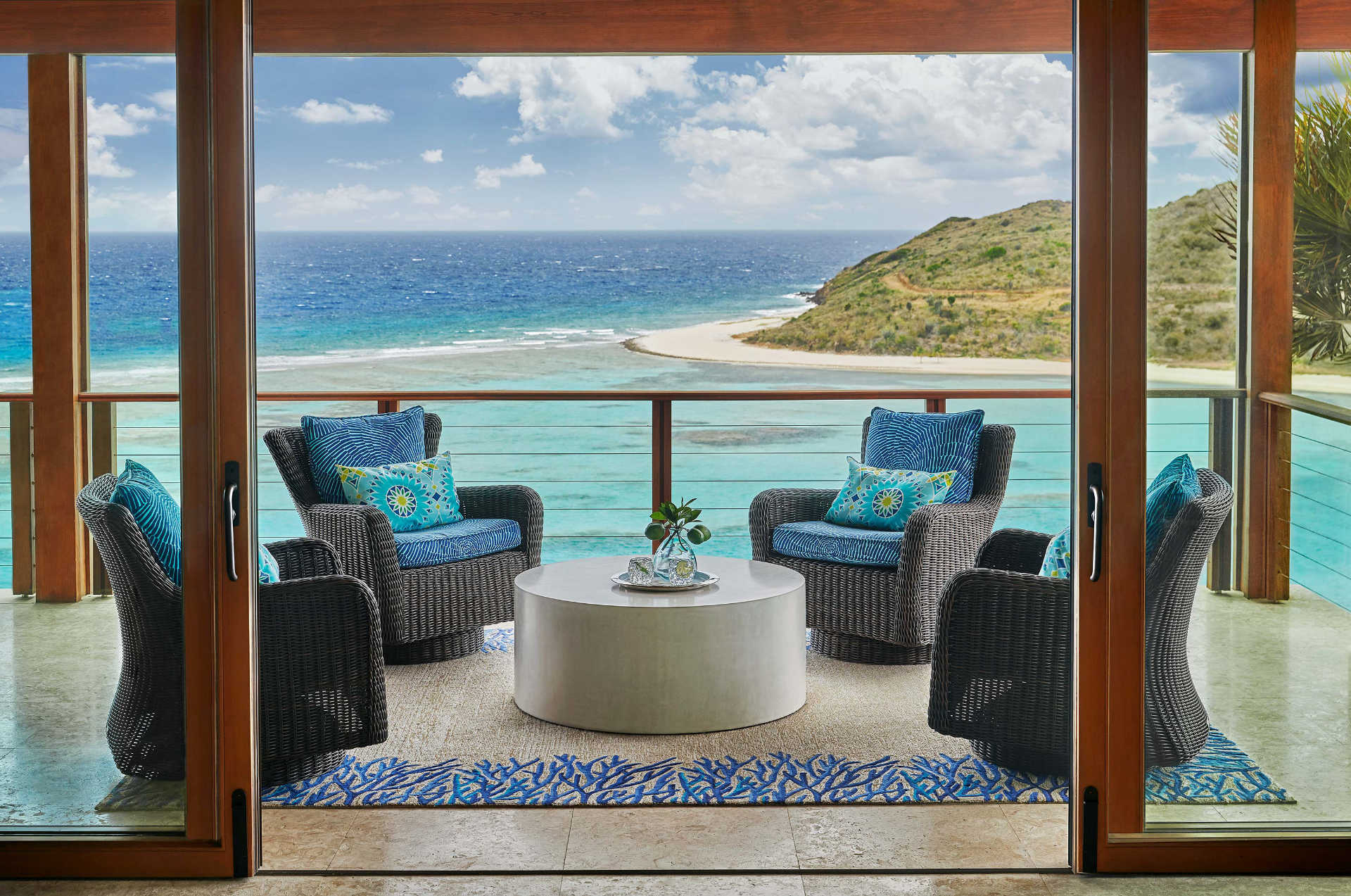 A cozy outdoor terrace at Poseidon's Perch overlooks an expansive ocean view. The area is furnished with wicker armchairs featuring blue cushions and patterned pillows, arranged around a white circular table. A blue rug with a coral motif adds a touch of coastal decor to this waterfront palace.