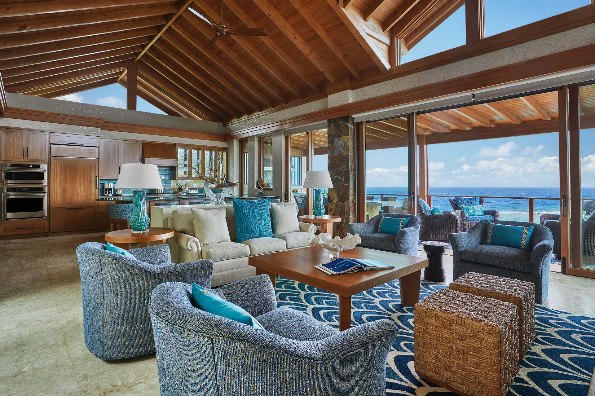 A spacious living room with wooden vaulted ceilings and large windows offering an ocean view in this waterfront palace called Poseidon's Perch. The room features blue armchairs, a beige sofa with turquoise cushions, and a wooden coffee table. The kitchen area includes wooden cabinets and modern appliances.