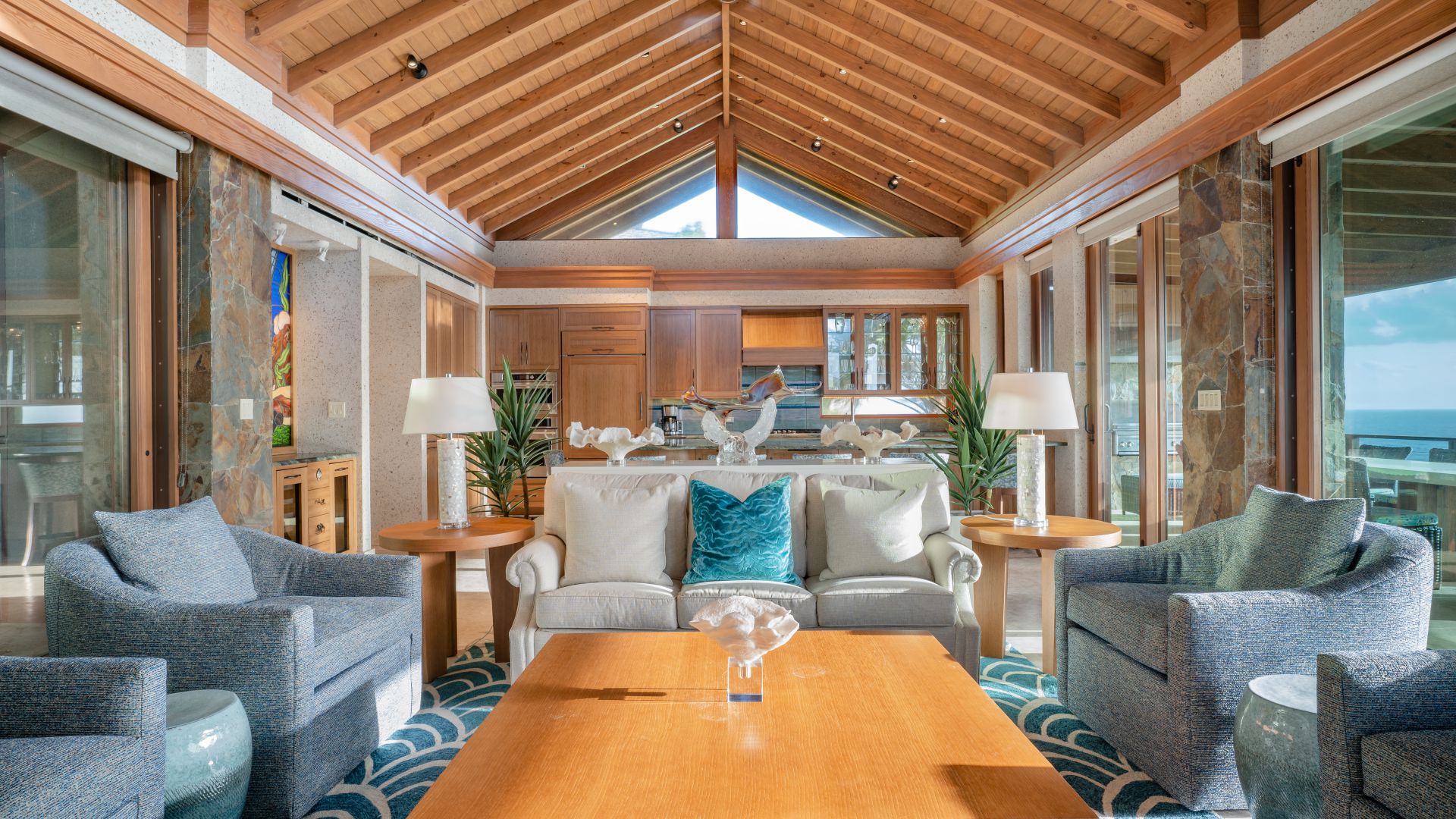 A luxurious living room in Poseidon's Perch, with a wooden vaulted ceiling, features large windows allowing abundant natural light. The room is elegantly furnished with blue armchairs, a beige sofa, and a wooden coffee table. The modern kitchen is visible in the background of this waterfront palace.