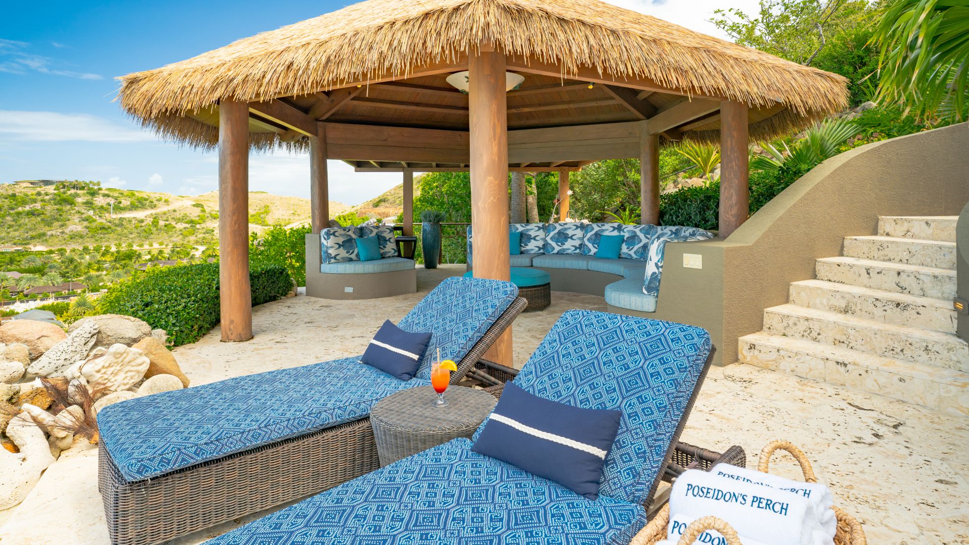A tropical outdoor lounge, aptly named Poseidon's Perch, features two blue-cushioned recliners with pillows and a wicker table holding a cocktail. Behind them, a thatched-roof pavilion houses a curved seating area with blue cushions. A staircase ascends to the right, lined with lush greenery.