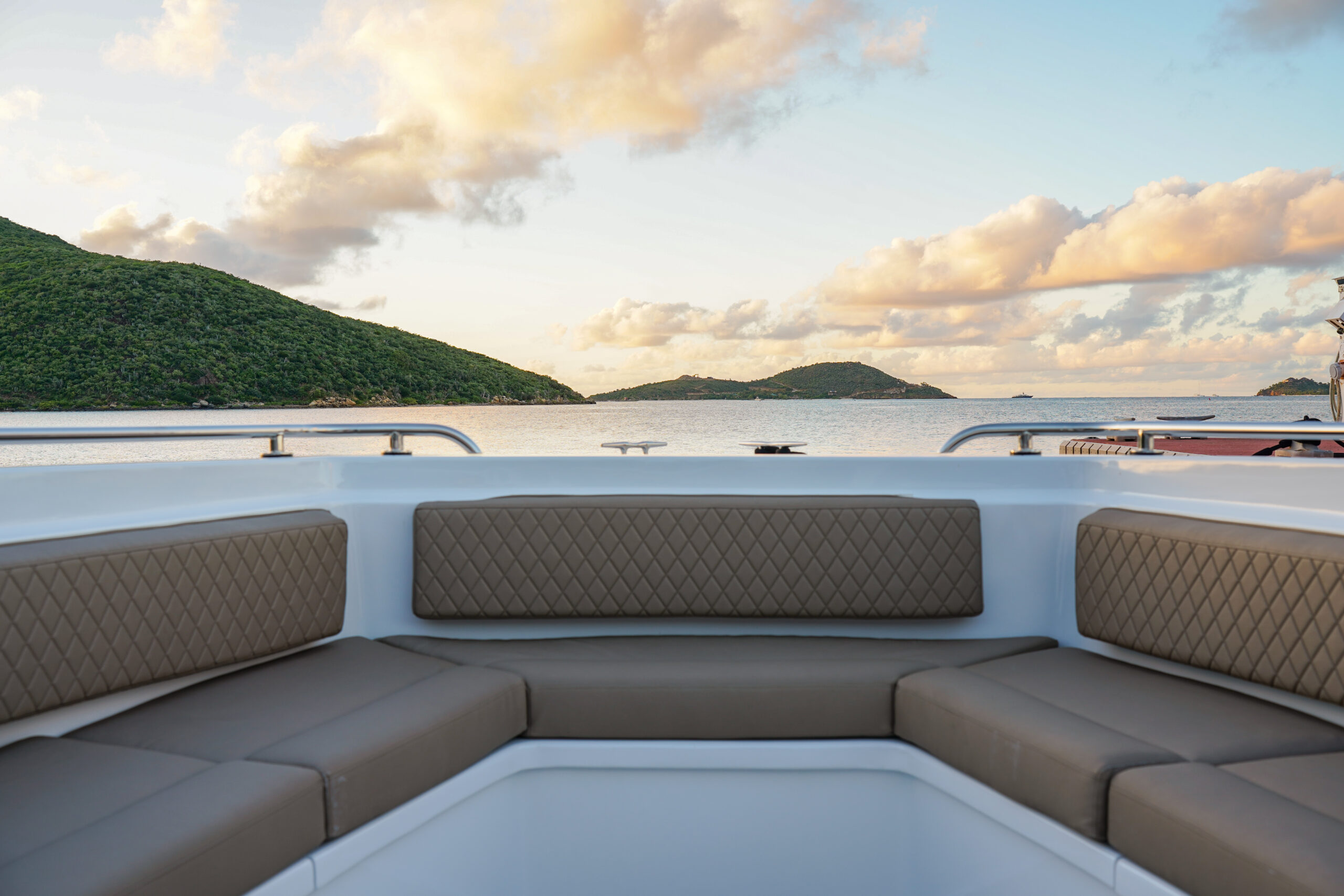 A view from the deck seating area of a boat, showcasing a tranquil scene of calm water and distant green islands under a sky with scattered clouds at sunset. The boat's cushioned brown upholstery boasts a modern design, inviting you to sail into serenity near Oil Nut Bay.