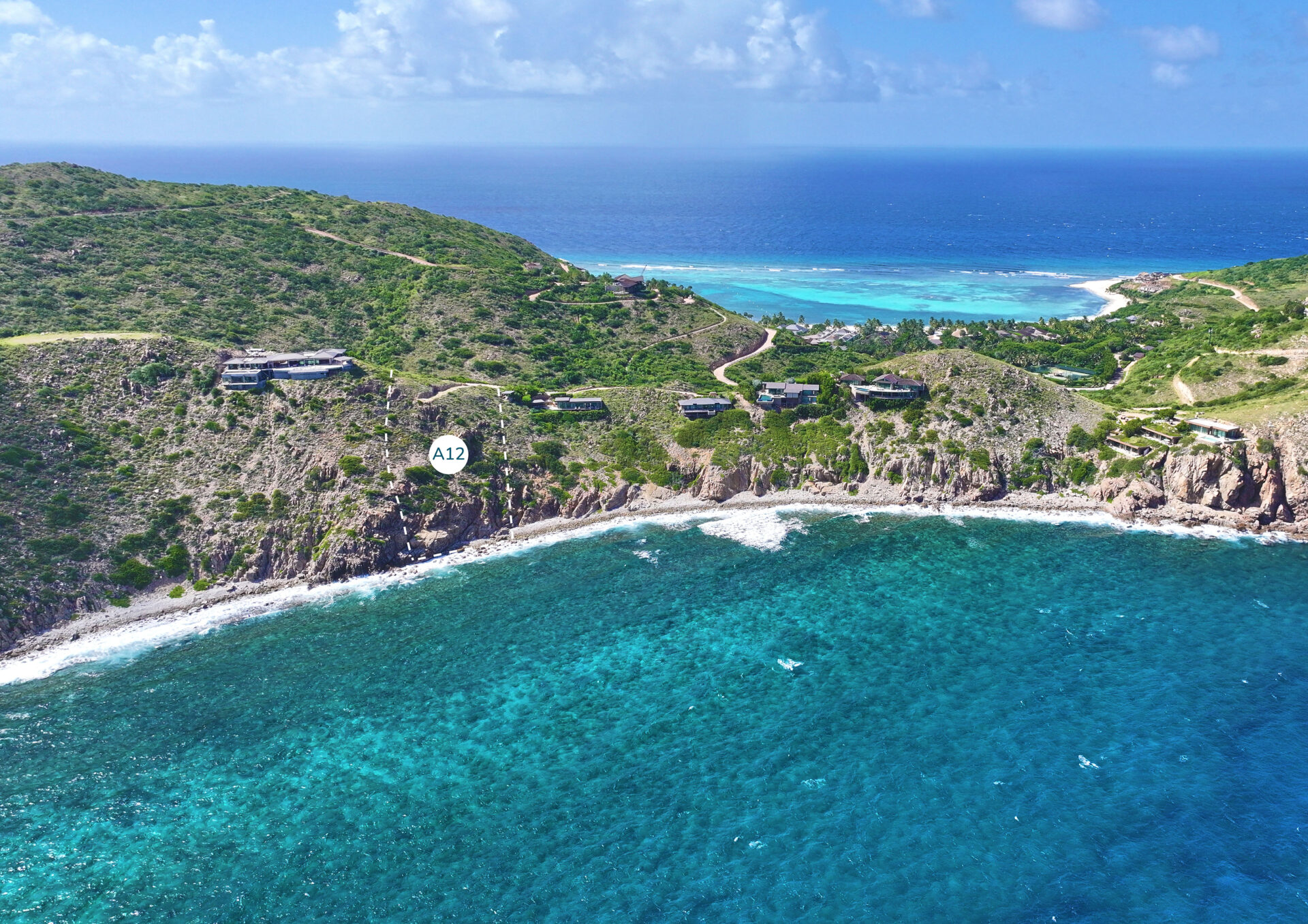 Aerial view of a coastal area with turquoise waters, rocky cliffs, and lush greenery. Several homesites are scattered on the cliffs overlooking the Atlantic Ridge. A white marker labeled "A12" is placed on one of the cliffs. The horizon shows a clear, sunny sky.
