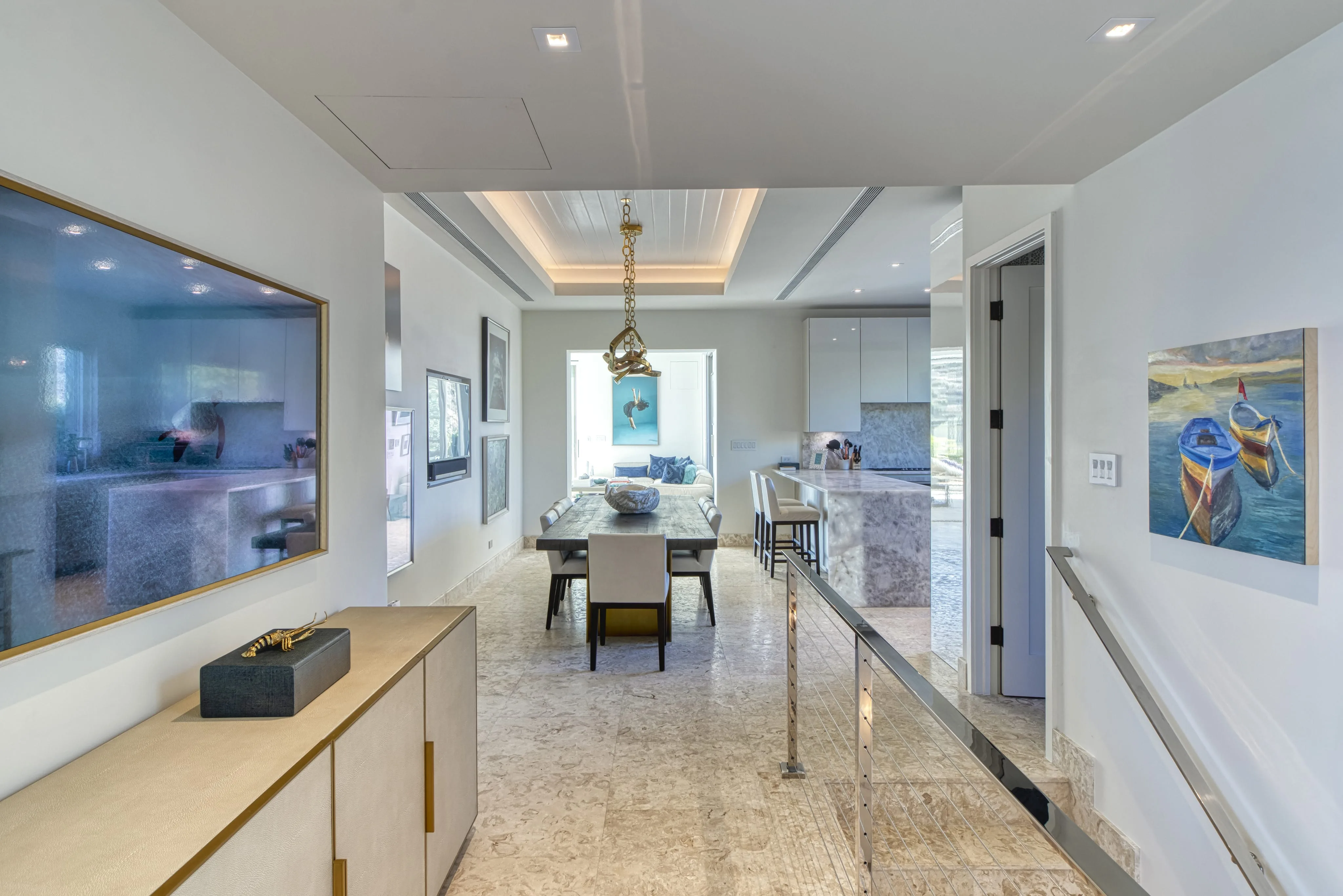 The modern dining room at Wild Side Villa boasts a rectangular dining table with eight chairs. It features a large photo on the left wall and a framed painting of two boats on the right. The space is completed with a light-colored marble floor and recessed lighting in the ceiling.