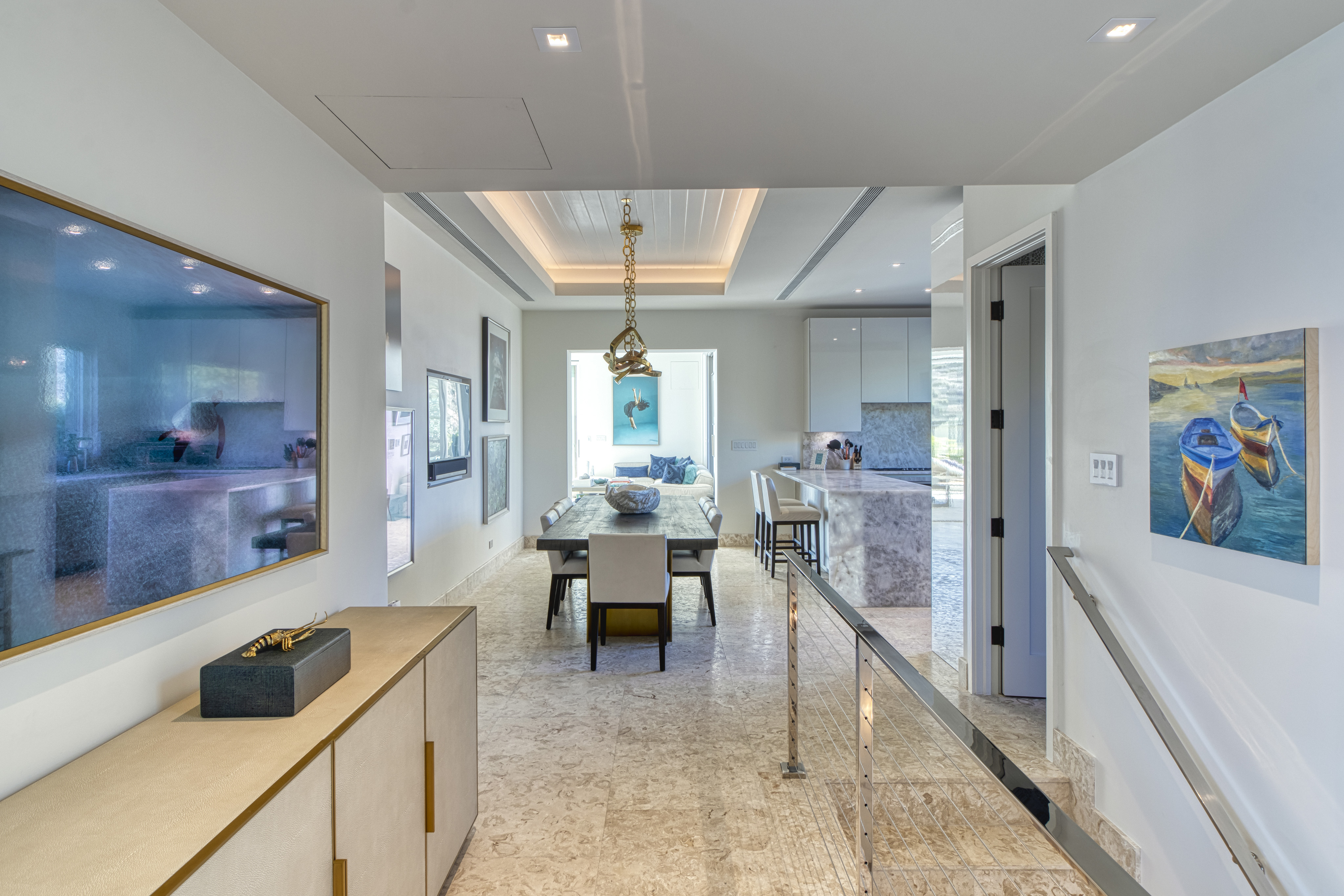 The modern dining room at Wild Side Villa boasts a rectangular dining table with eight chairs. It features a large photo on the left wall and a framed painting of two boats on the right. The space is completed with a light-colored marble floor and recessed lighting in the ceiling.