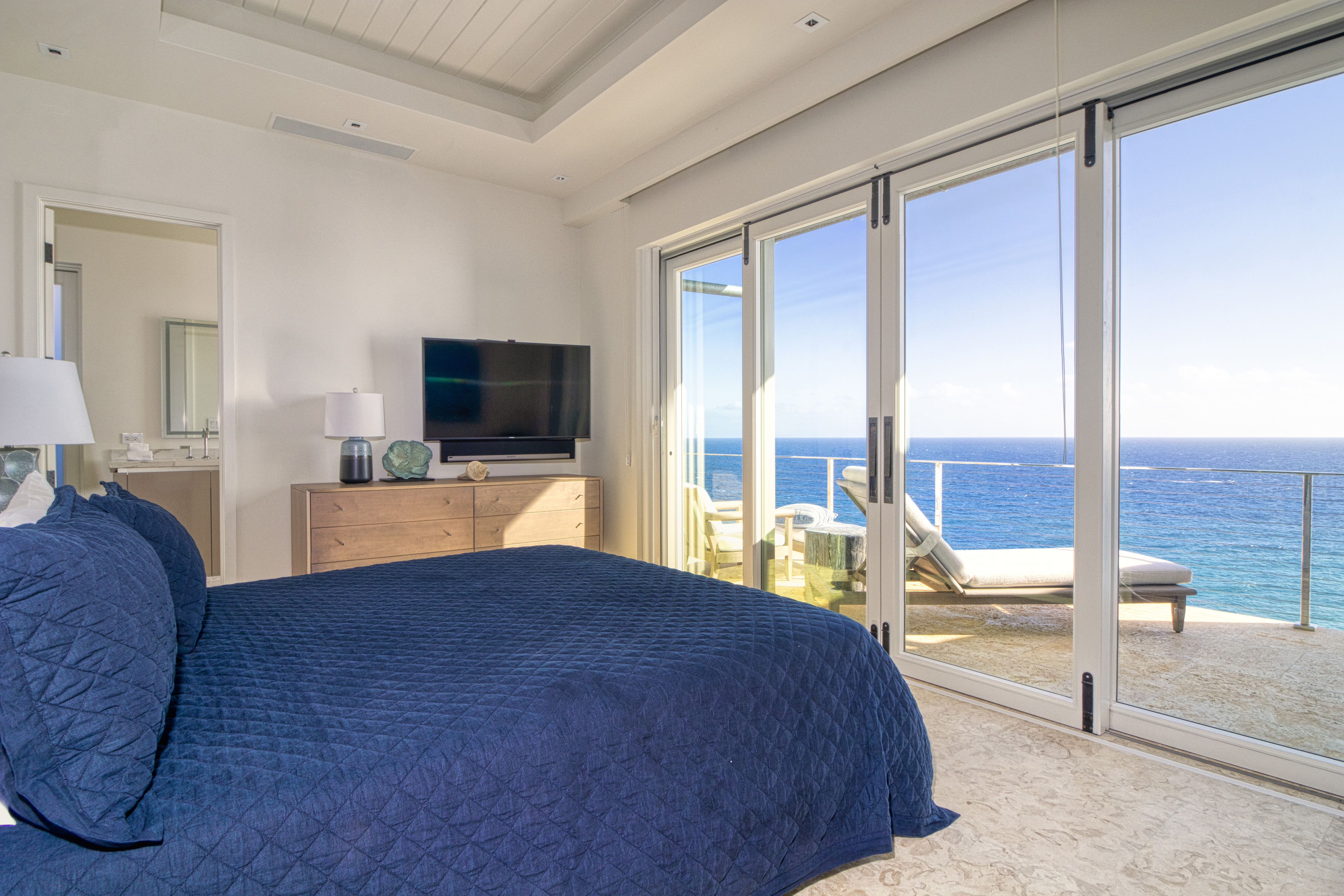A modern bedroom at Wild Side Villa features a queen-sized bed with a dark blue bedspread facing a flat-screen TV mounted on a wooden dresser. Large sliding glass doors open to a balcony with sun loungers, offering a stunning ocean view. Light beige walls and floor complement the airy space.