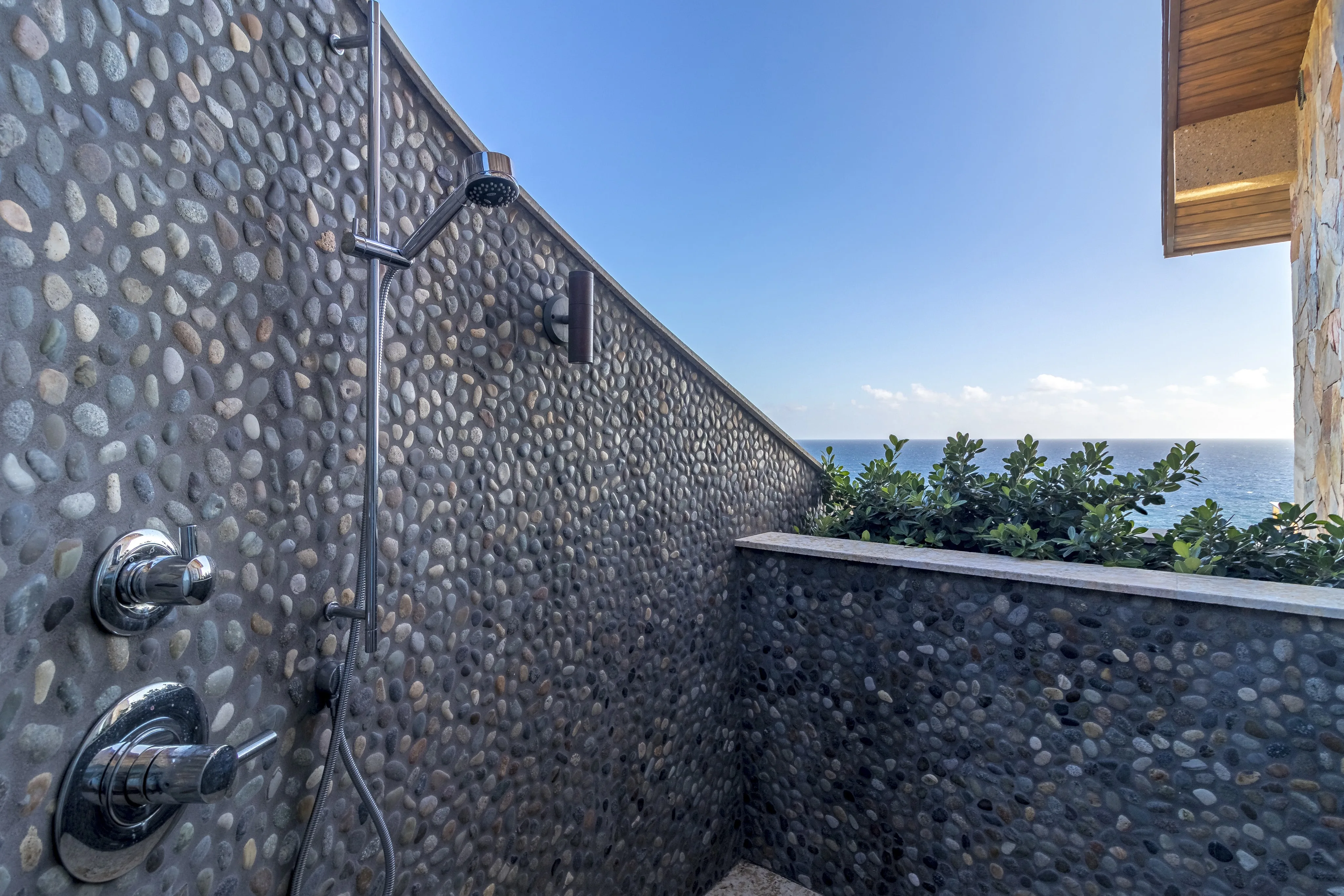Outdoor stone shower with modern metal fixtures at Wild Side Villa, overlooking a scenic ocean view under a clear blue sky. Lush green plants and a stone building edge partially visible on the right side.