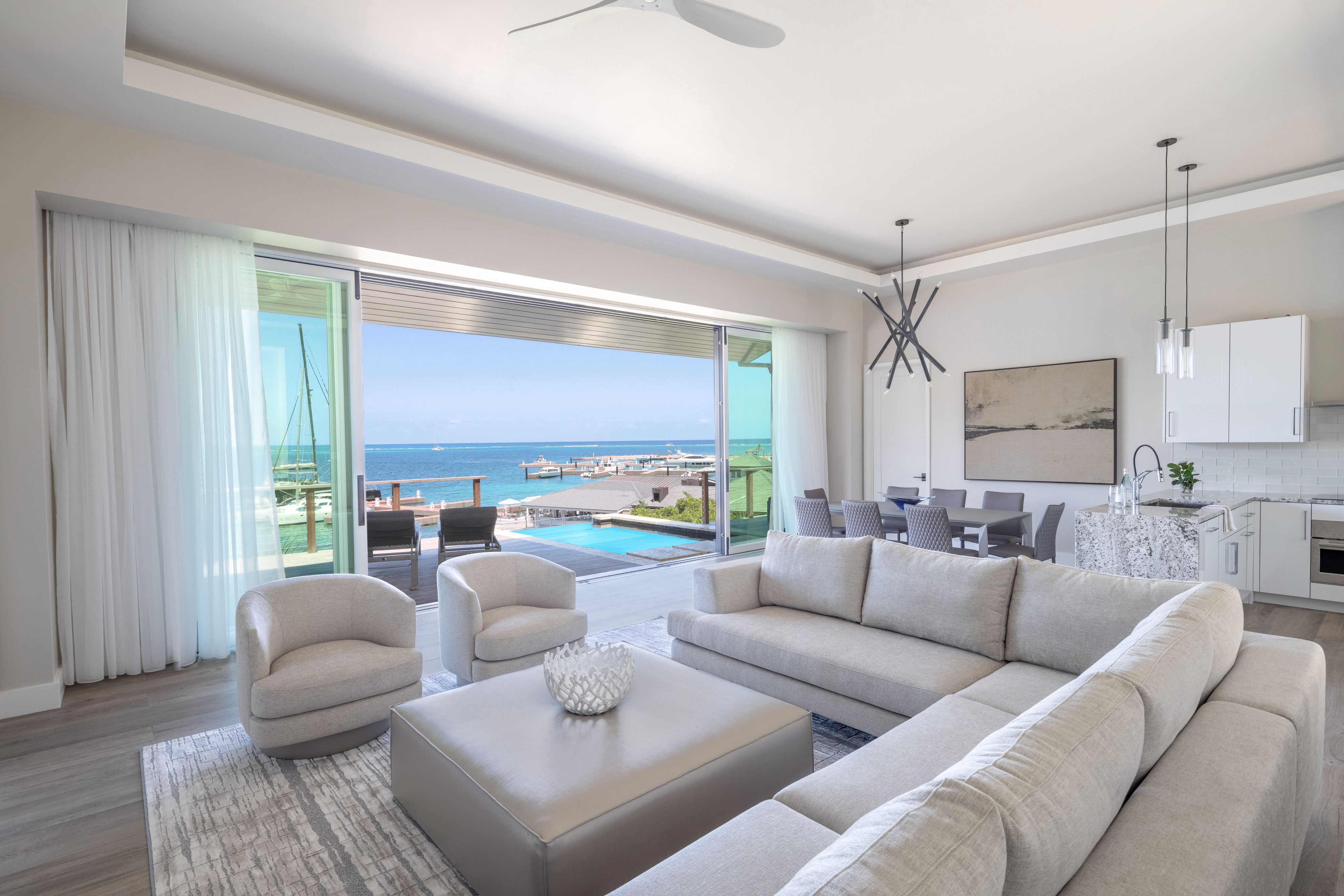 A spacious, modern living room with a sectional sofa, two armchairs, and a large ottoman. Floor-to-ceiling glass doors open to a patio with a pool, offering an ocean view. This elegant villa's open-concept area includes a dining table and a contemporary kitchen perfect for enjoying an Amarone.