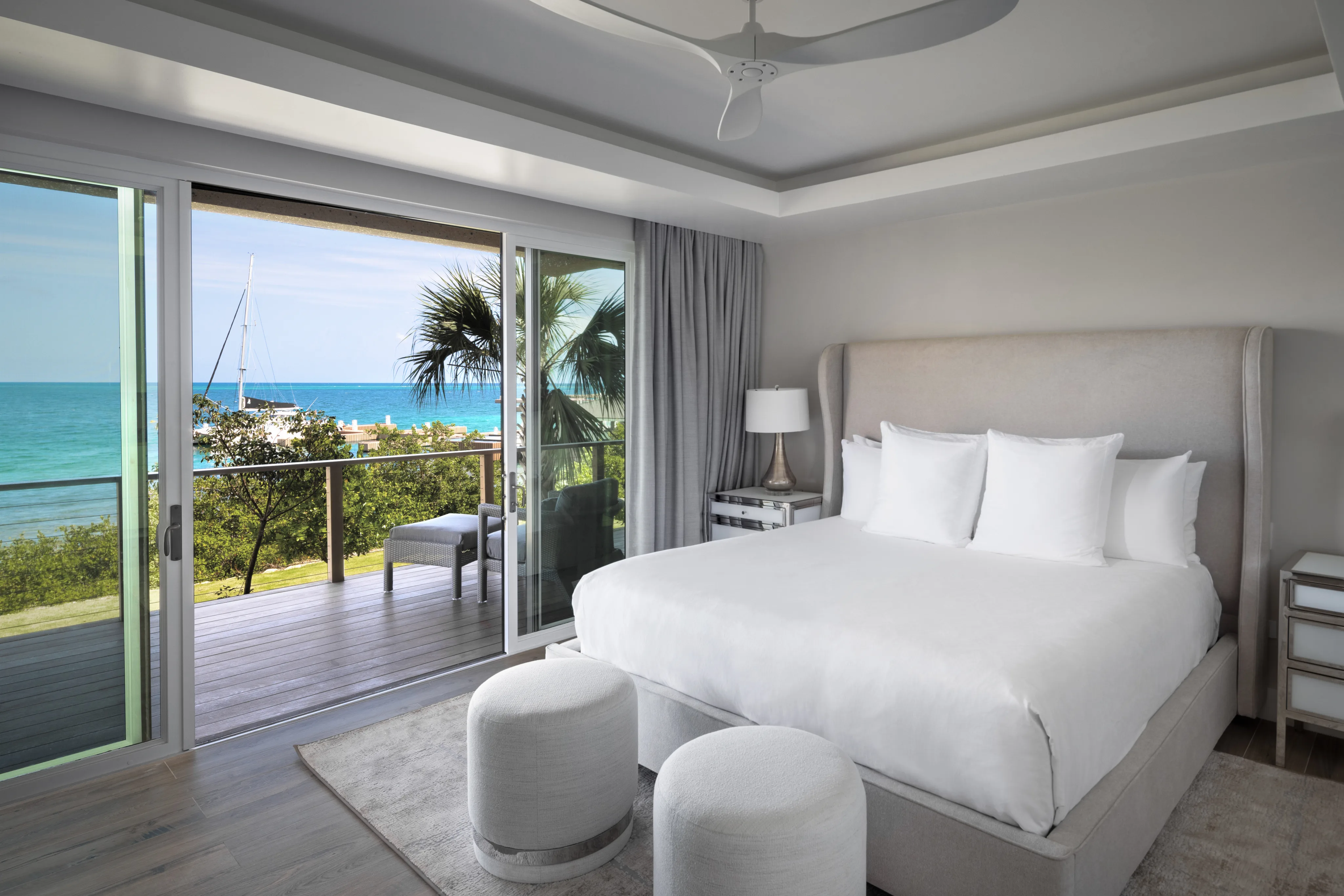 Modern villa bedroom with large sliding glass doors offering a scenic ocean view. The room features a plush bed with white linens, two round ottomans at the foot of the bed, and light-colored furniture. A balcony with a palm tree and sailboats can be seen outside.