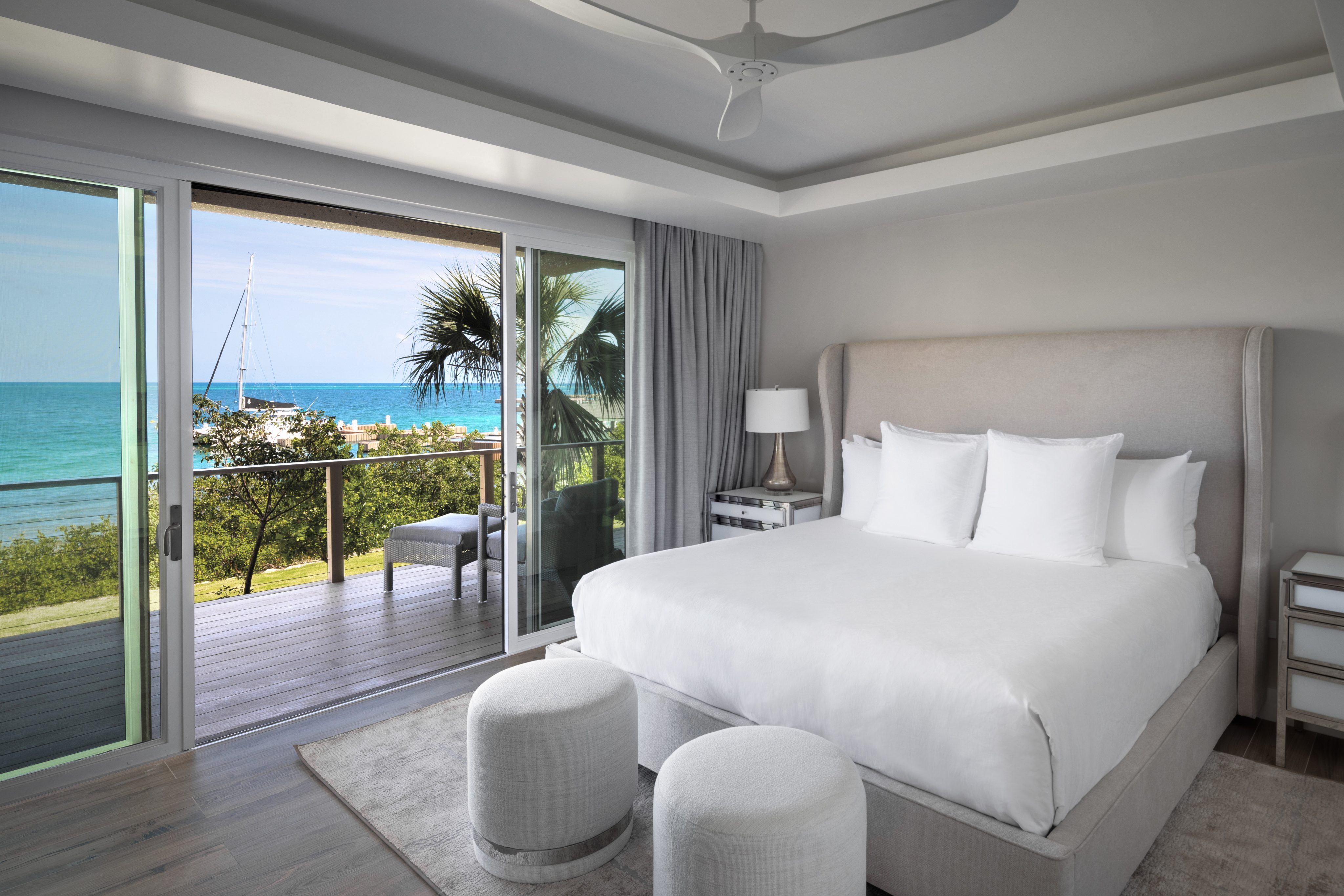 Modern villa bedroom with large sliding glass doors offering a scenic ocean view. The room features a plush bed with white linens, two round ottomans at the foot of the bed, and light-colored furniture. A balcony with a palm tree and sailboats can be seen outside.