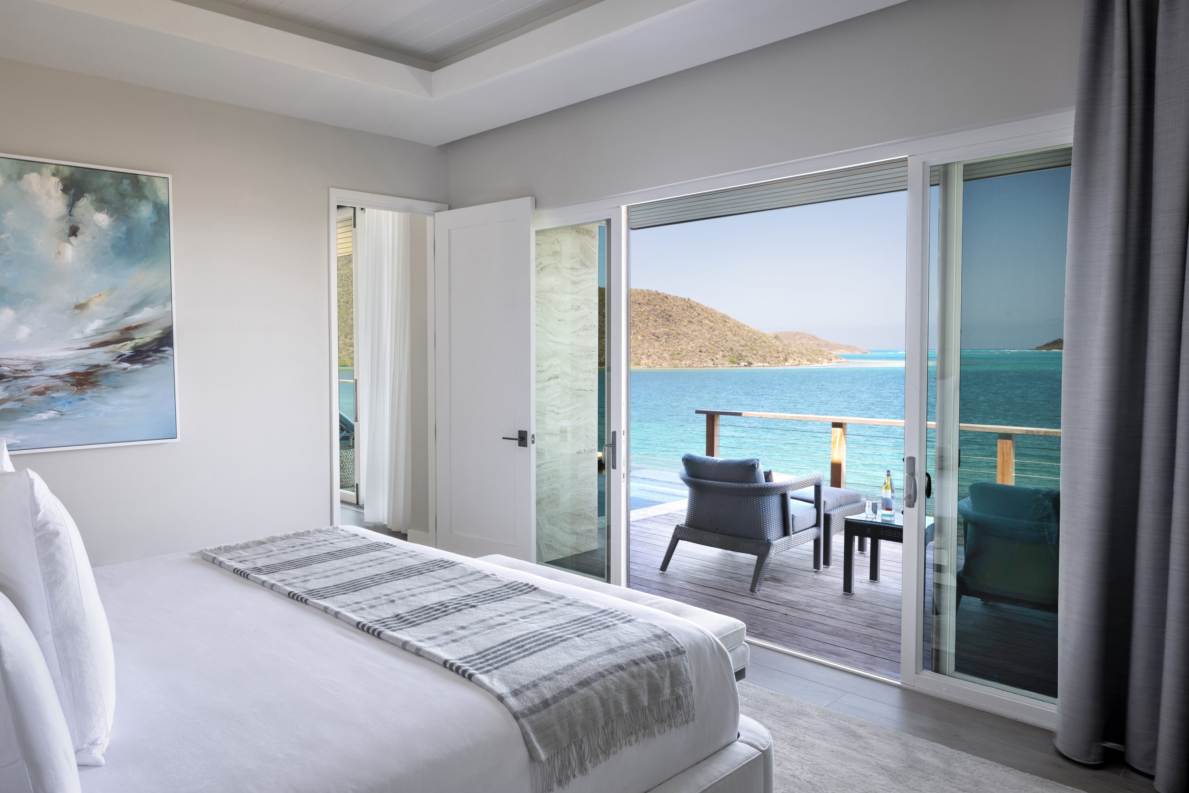 A modern bedroom in this villa features a neatly made bed with a striped throw blanket. Large sliding glass doors open to a wooden balcony with two lounge chairs, offering a scenic view of the calm, blue ocean and distant hills under a clear sky, perfect for sipping an Amarone.