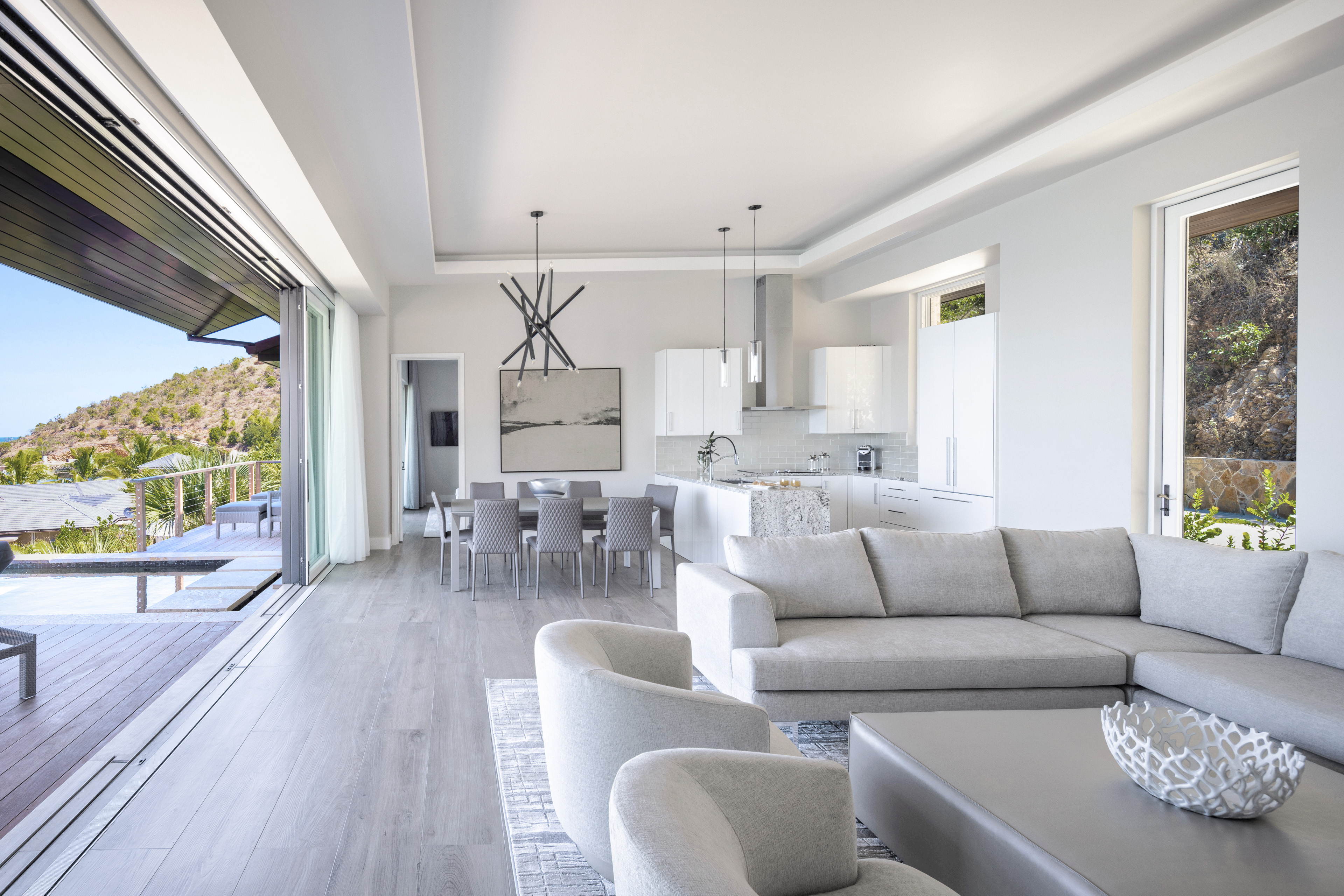 Modern, open-plan living and dining area with minimalist furniture in neutral tones. Large glass doors open to a deck with views of hills and greenery. Contemporary lighting fixtures hang above the dining table, and the kitchen in this exquisite villa features sleek white cabinetry.