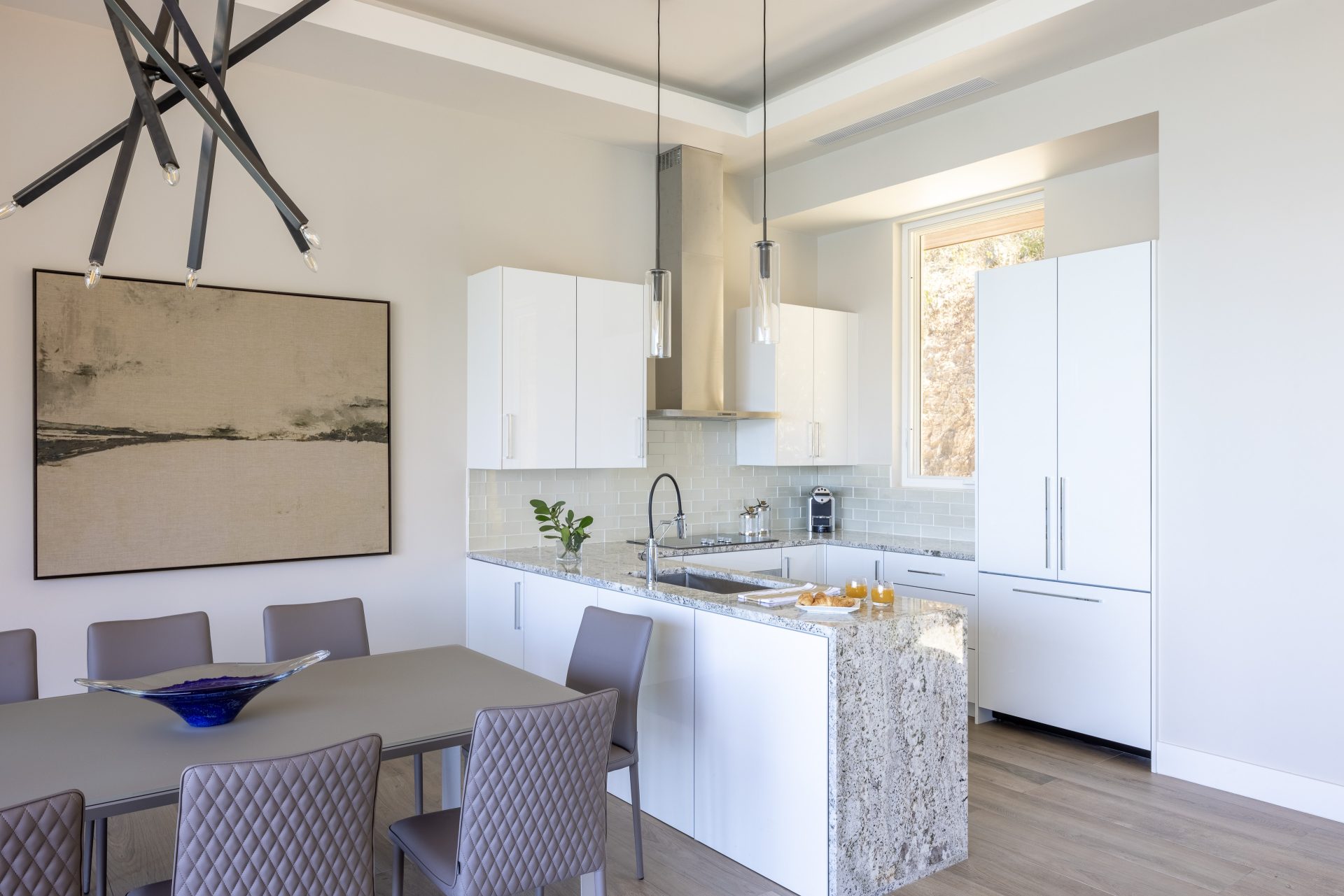 Modern kitchen and dining area in this villa feature light-colored walls and wooden flooring. Kitchen boasts white cabinets, granite countertops, a central island with a sink, and stainless-steel appliances. Dining area includes an Amarone-hued table with matching quilted chairs. Large abstract painting and unique black chandelier hang nearby.