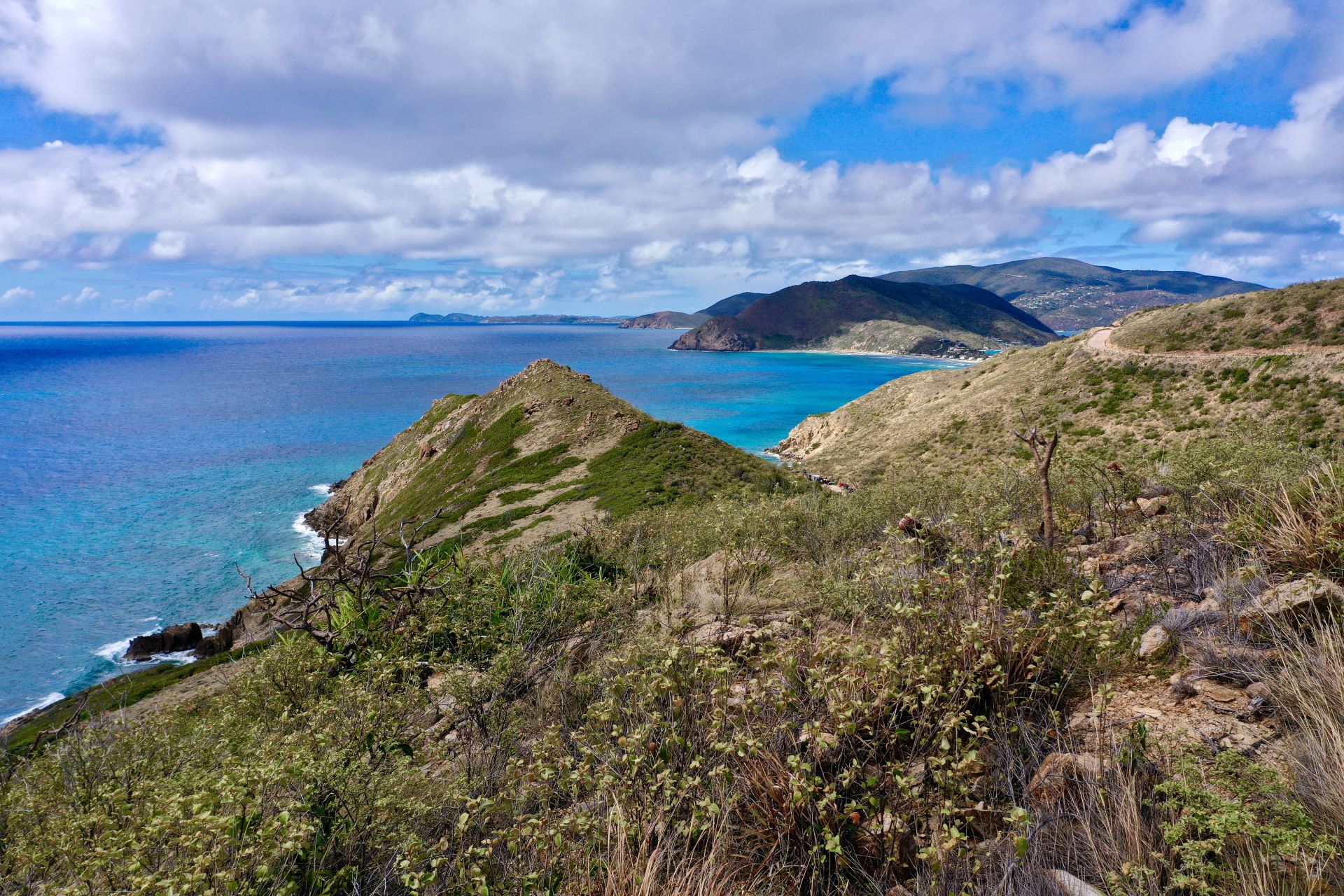 A scenic view of a coastal landscape featuring clear blue waters meeting rocky shores. Rolling hills covered in grass and shrubs extend towards the water. The sky is partly cloudy with patches of blue. The foreground includes dry, rugged terrain with sparse vegetation near the Boulder Homesite.