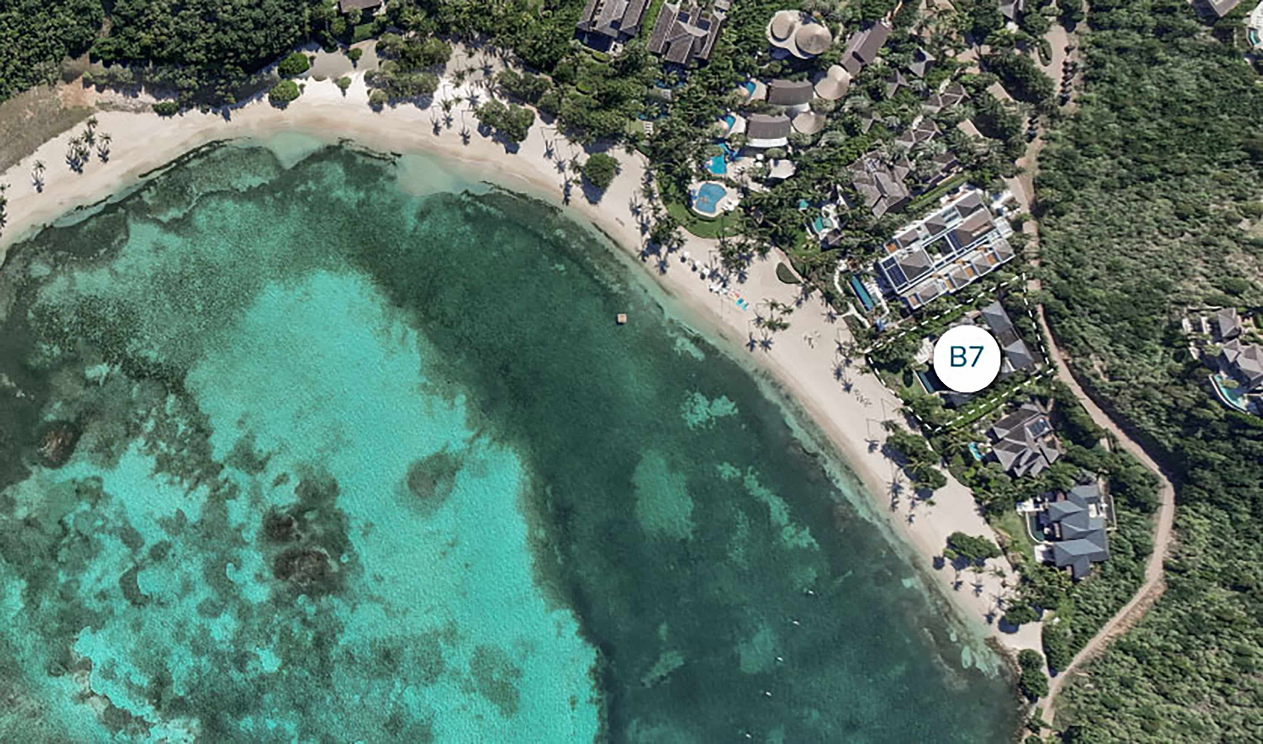 An aerial view of a tropical beachfront resort, featuring clear turquoise waters and white sandy beaches. The resort, which includes multiple buildings and pools surrounded by lush greenery, also offers charming beach houses. A tag labeled "B7" is positioned near one of the buildings.