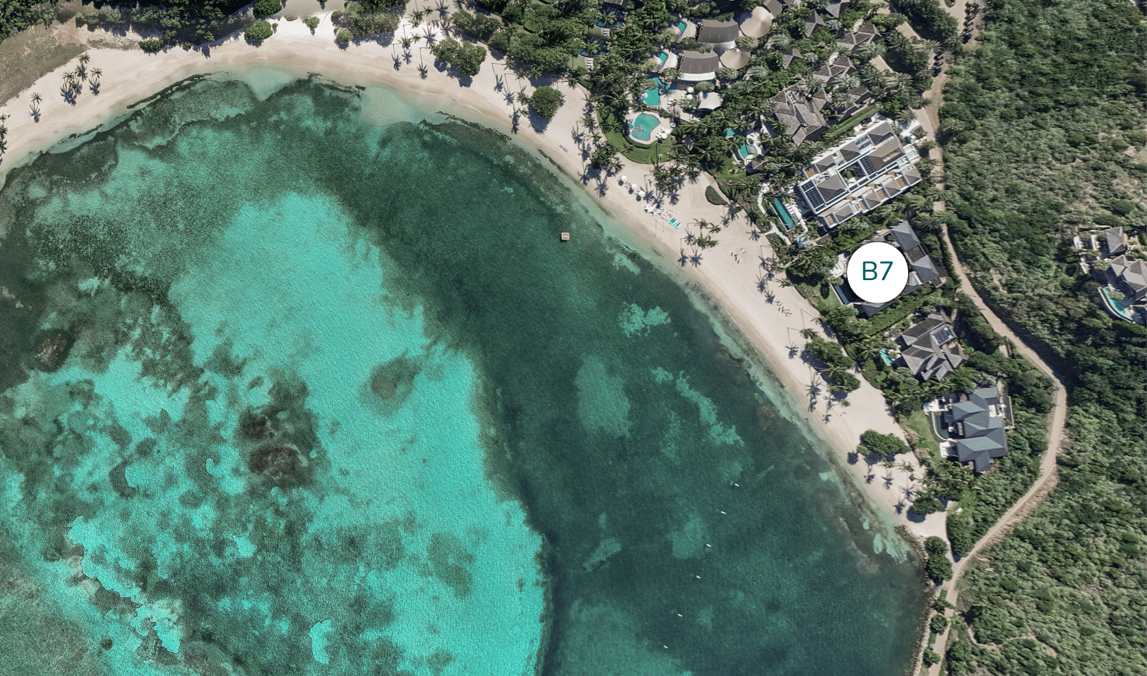 Aerial view of a coastal area with turquoise waters and a sandy beach. Luxurious villas, including the exquisite Cheemaun Villa, are nestled among greenery along the shoreline. A digital marker labeled "B7" is positioned above one of the properties.