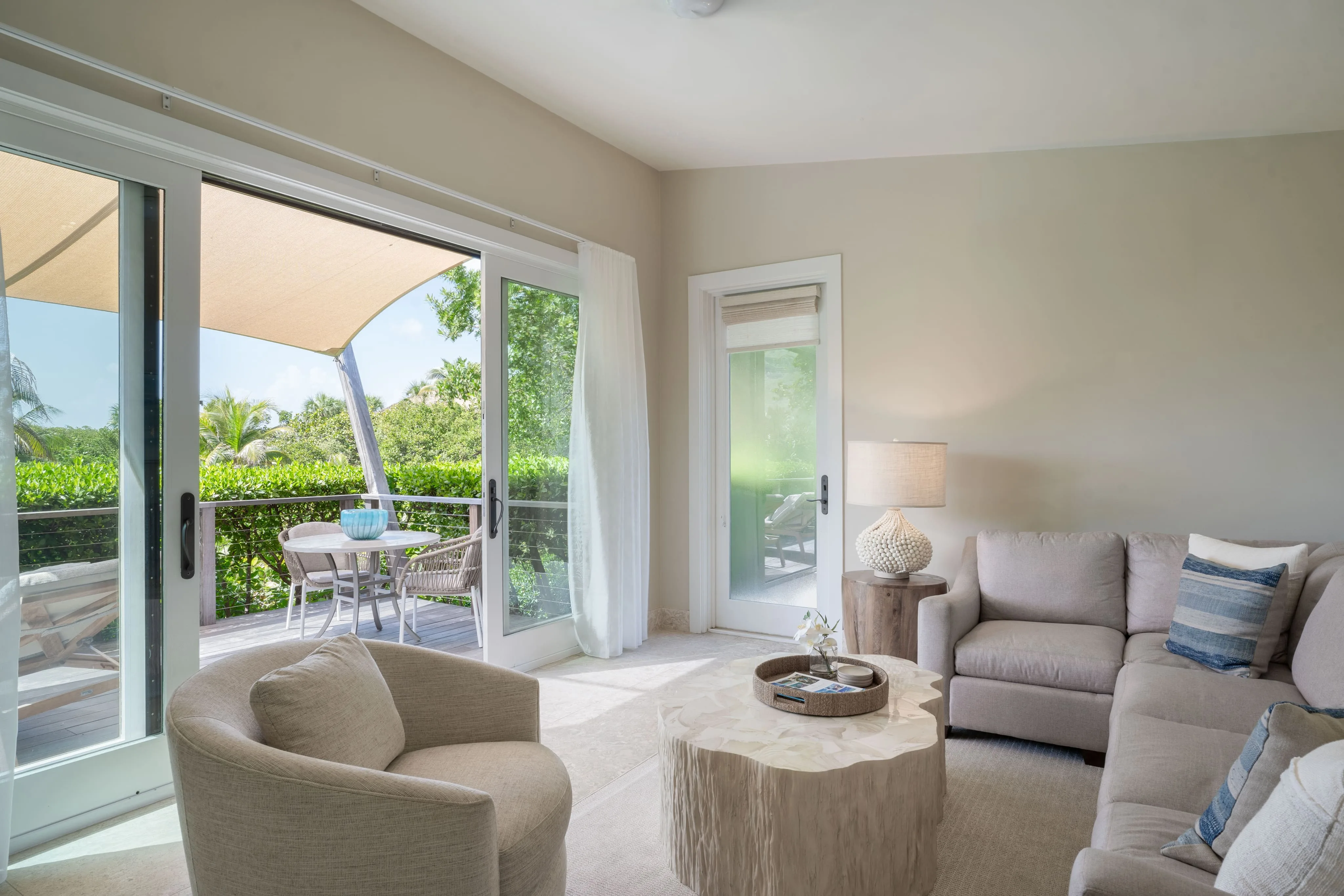 A bright, modern living room in the Bay Suites features a beige sectional sofa, a round armchair, and a wooden coffee table. Glass doors open to a patio with a canopy, outdoor seating, and lush greenery. Natural light fills the room, enhancing the serene and inviting atmosphere.
