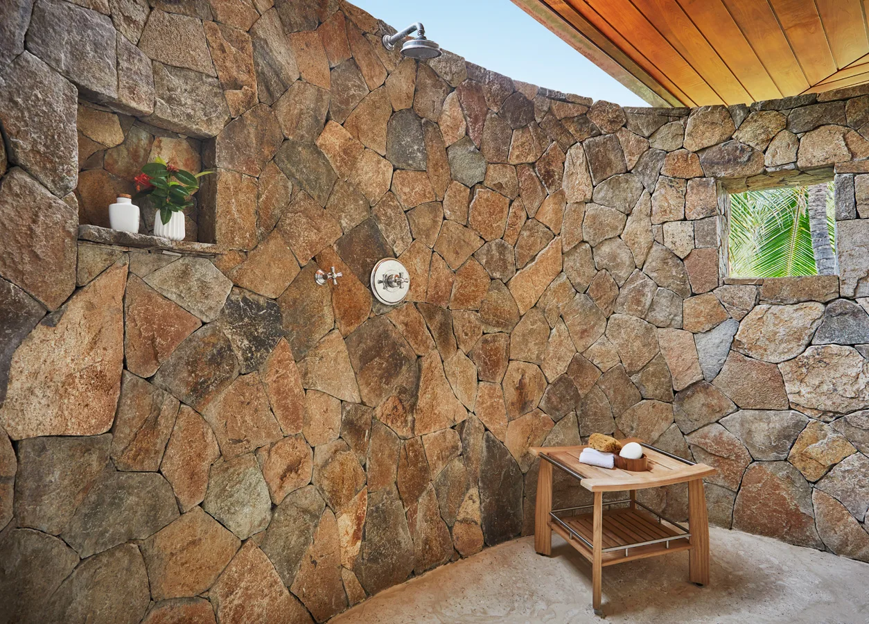 An outdoor stone shower area at the villa features a wall of large, irregularly shaped rocks, a wooden bench with folded towels and soap, and two niches holding a red potted plant and white jar. An overhead showerhead is mounted on the wall. A small window reveals greenery, enhancing the serene ambiance of Villa Cheemaun.