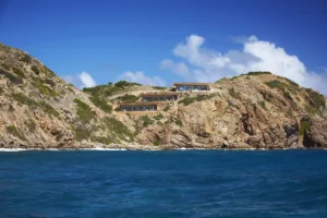 The Cliff Suites, a luxurious modern villa, sits perched on a rocky cliff overlooking a pristine blue ocean. Surrounded by rugged terrain and sparse vegetation, it offers panoramic views and blends seamlessly with the natural landscape under a clear, vibrant sky.